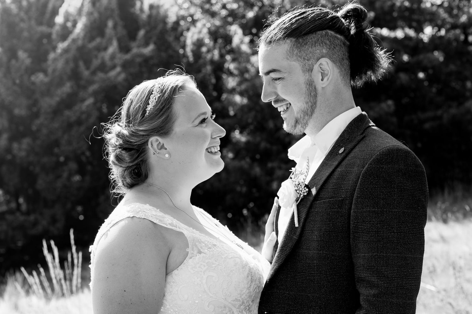 Black and white portrait of smiling bride and groom with man bun hairstyle at outdoor wedding.