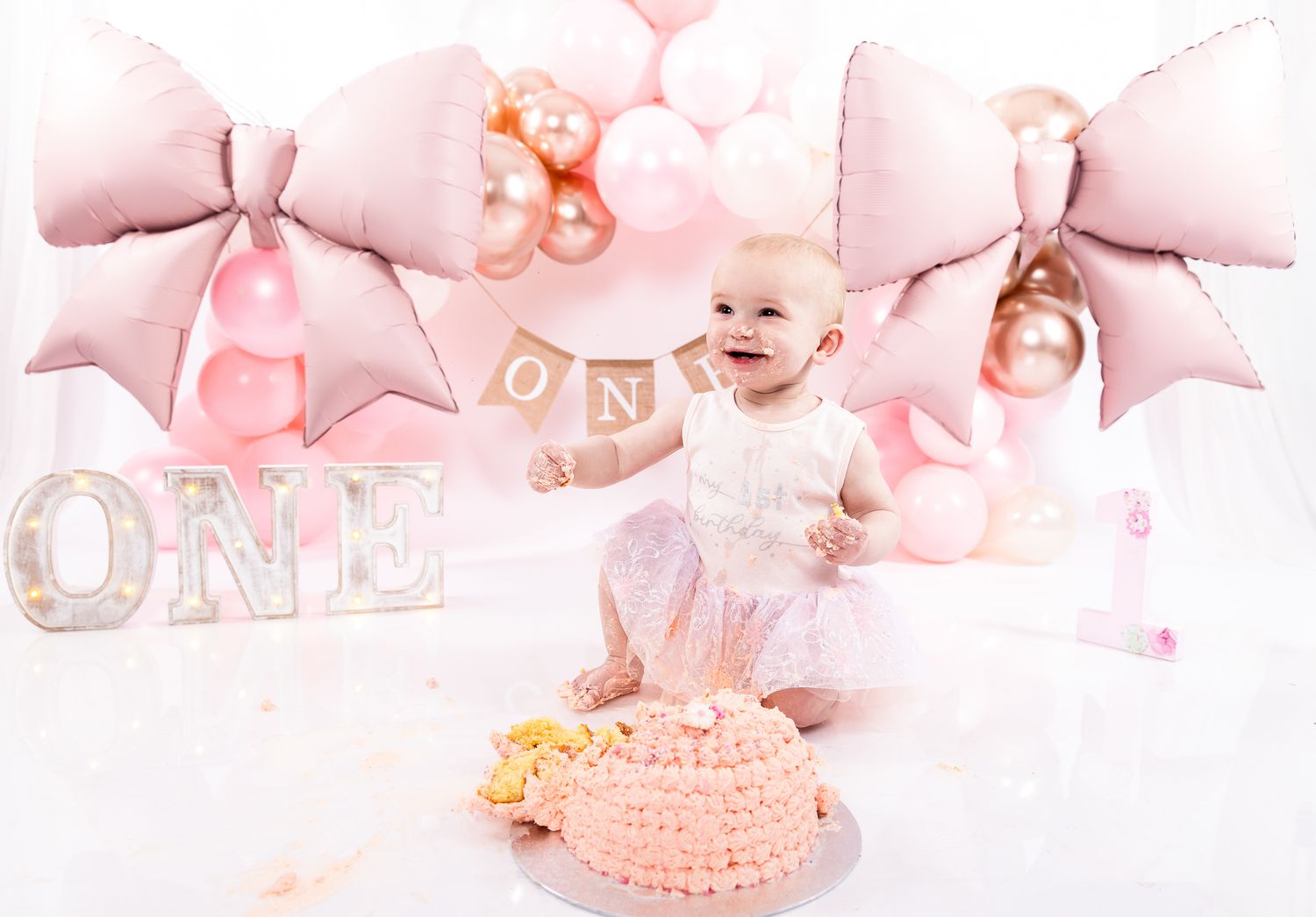 Baby girl celebrating first birthday with pink cake, ONE letters, bow balloons, and pastel balloon garland backdrop.