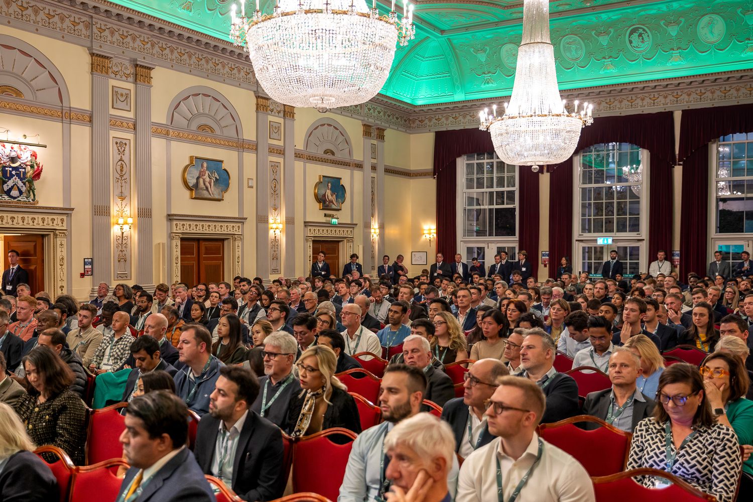 Large audience seated in an ornate conference hall with chandeliers during a business summit.