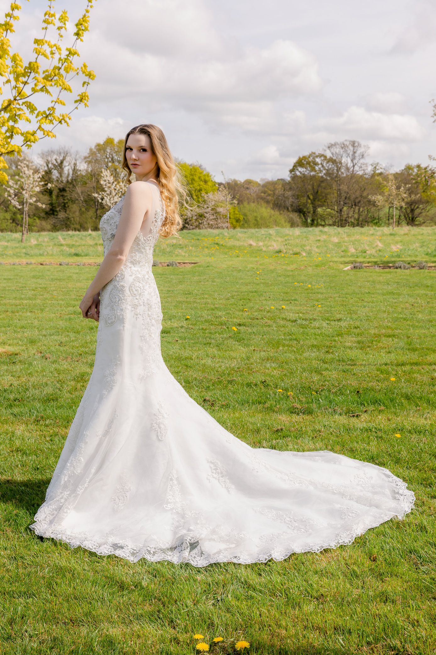 Elegant bride in lace mermaid wedding gown with train posing on green grass in outdoor setting.