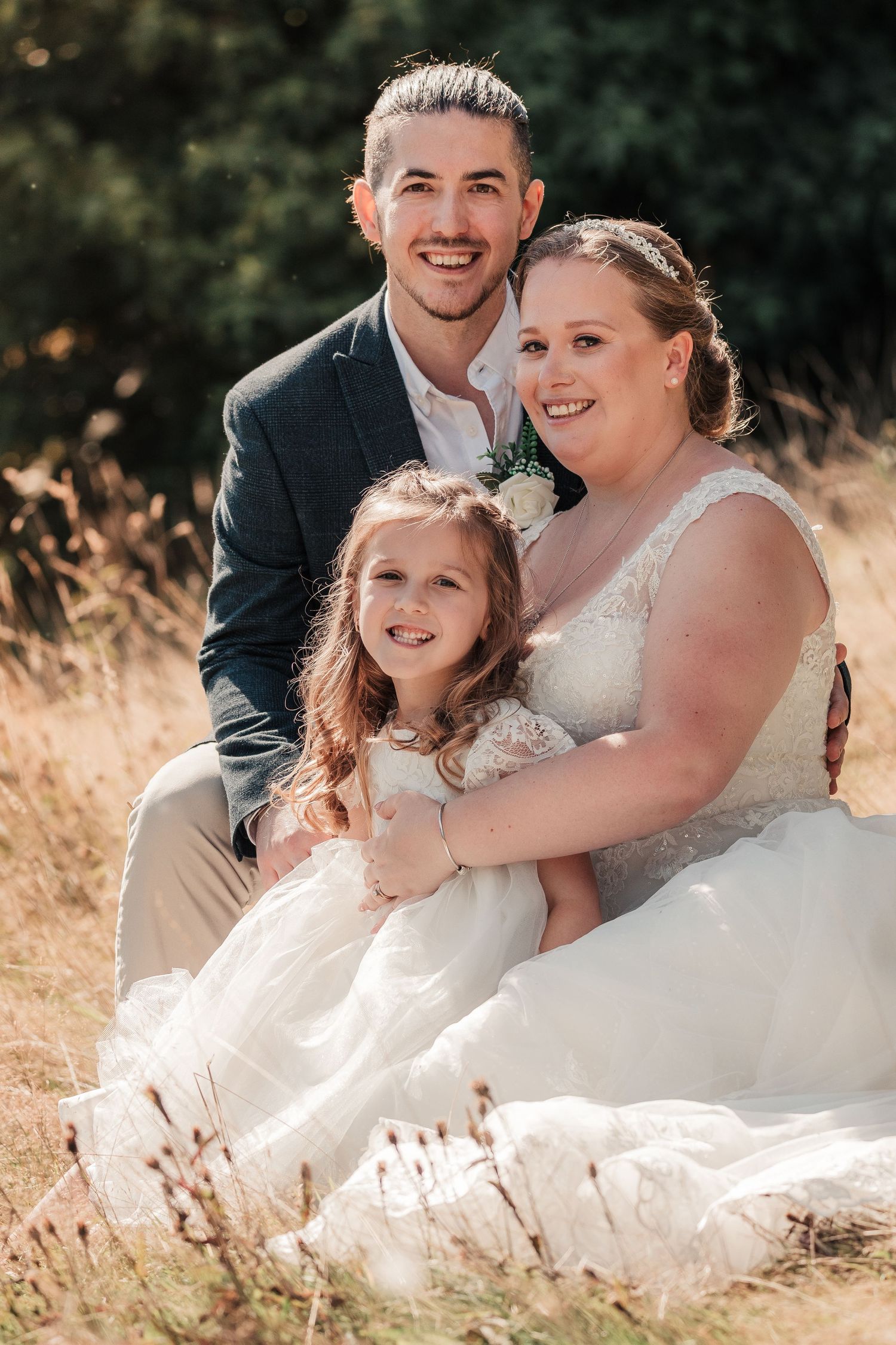 Smiling bride and groom sitting in a sunny field with a young flower girl in a white dress.