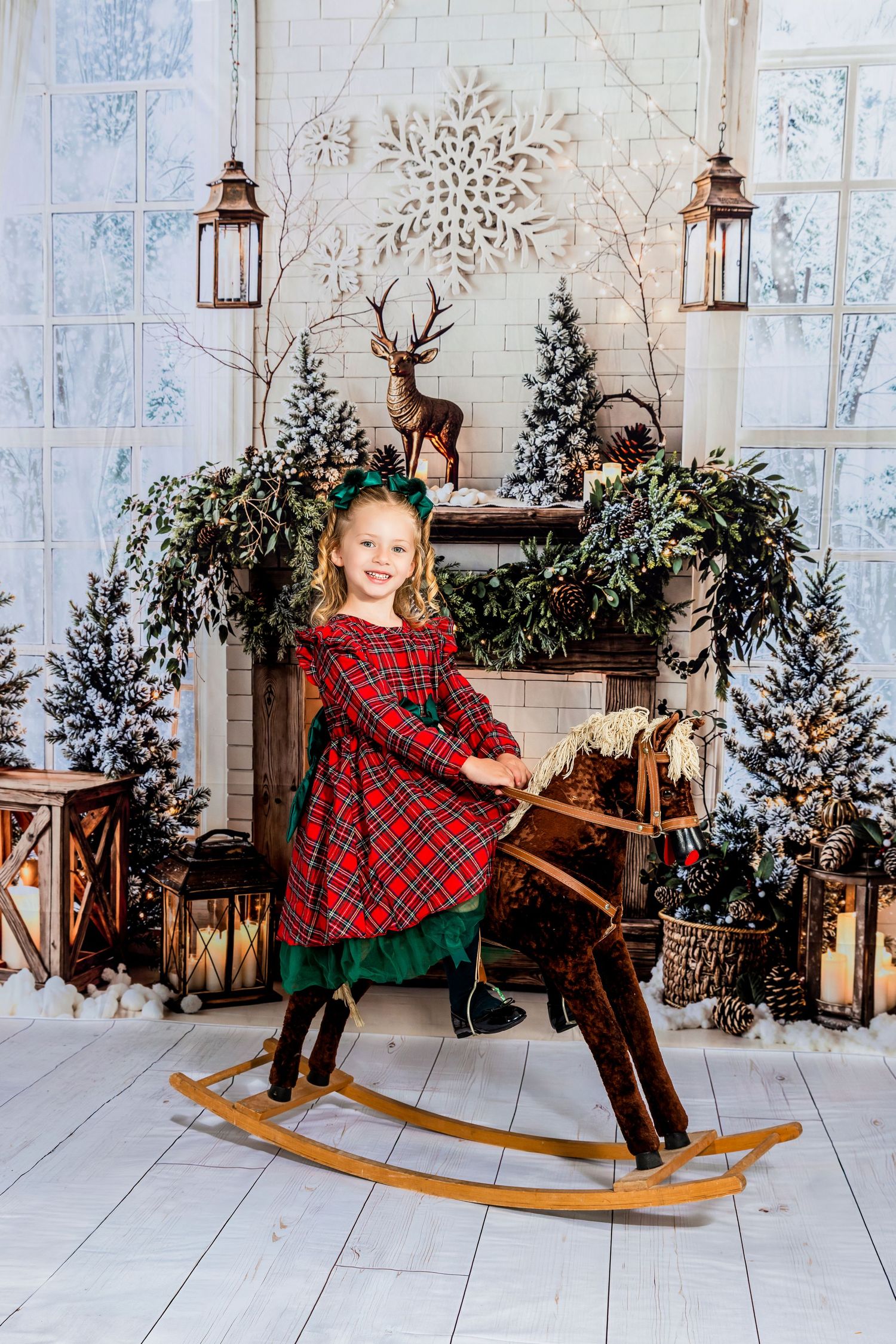 Young girl in red plaid dress riding a wooden rocking horse in a festive Christmas decorated studio setting.