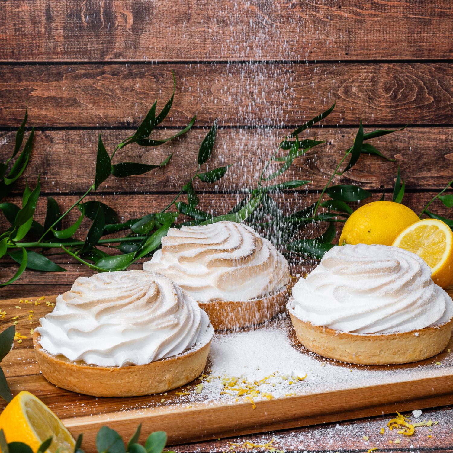 Three lemon meringue tarts dusted with powdered sugar on a wooden board with fresh lemons.