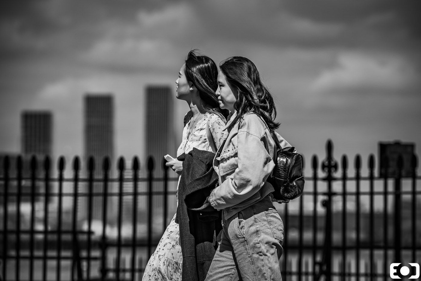 Two young women walking together against city skyline backdrop, black and white street photography.