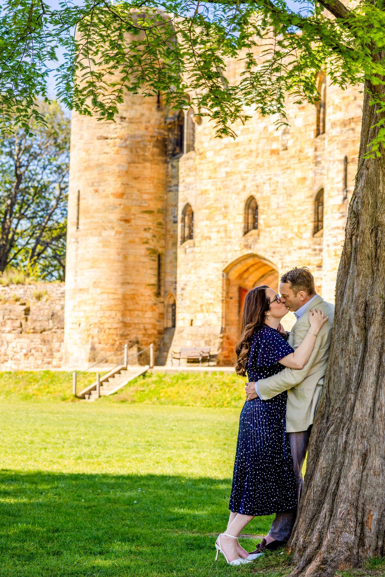 Couple kissing by a tree with a historic stone castle in the background on a sunny day.