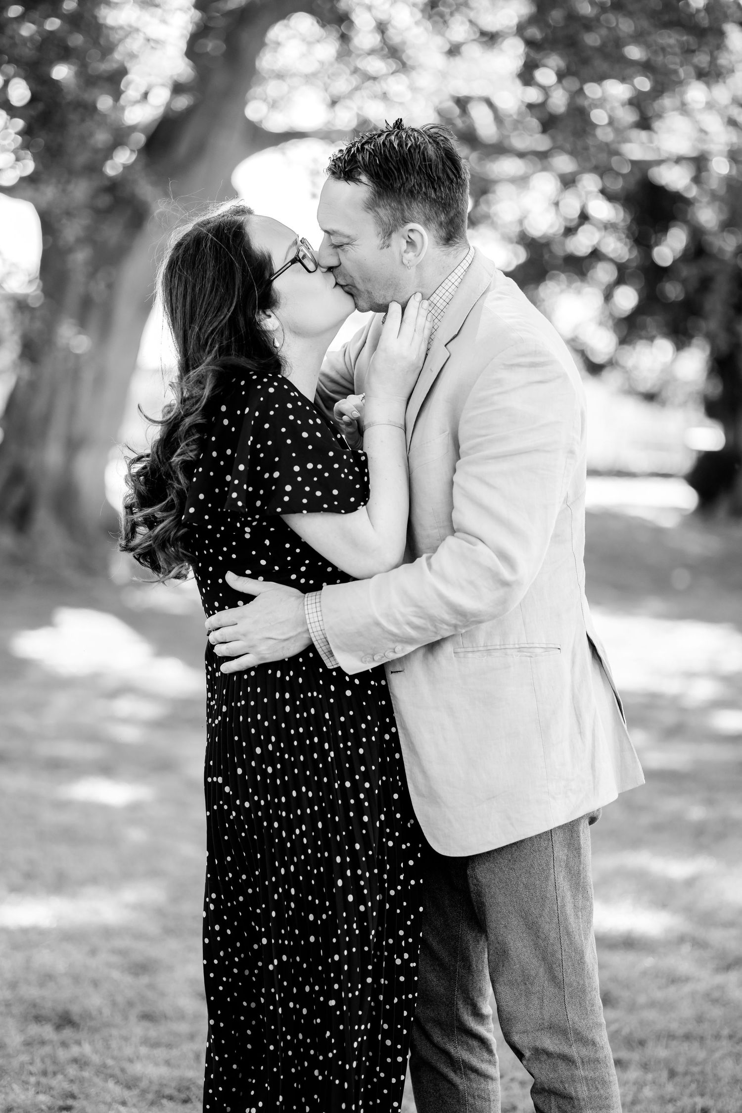Romantic black and white photo of a couple kissing in a sunlit park setting.