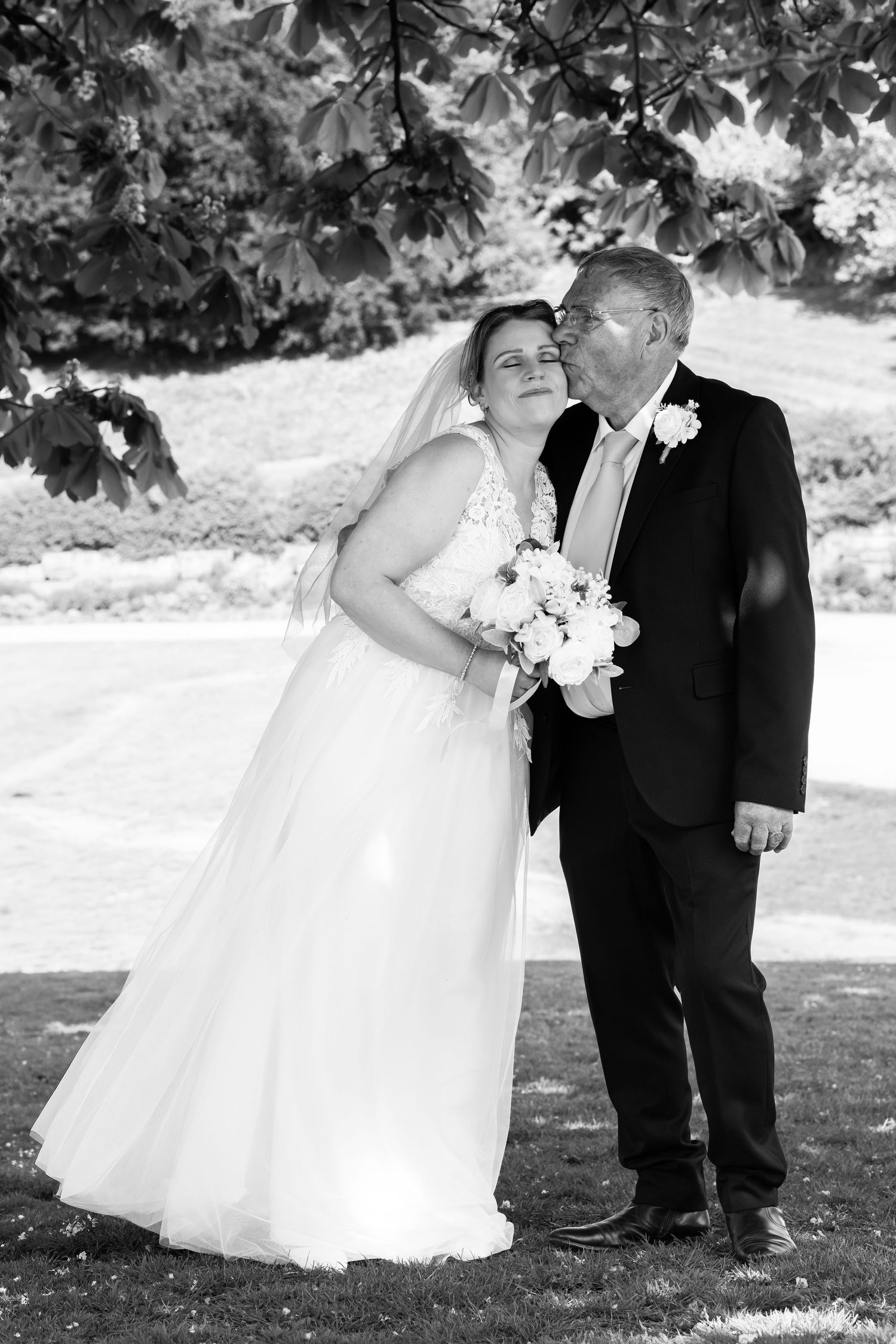 Black and white photo of a groom kissing bride on cheek outdoors, bride holding white bouquet in wedding dress with veil.