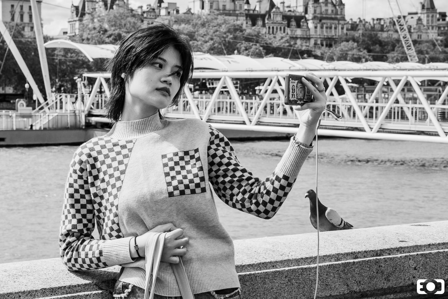 Young woman in checkered sweater feeding a pigeon by the Thames River in London, black and white street photography.