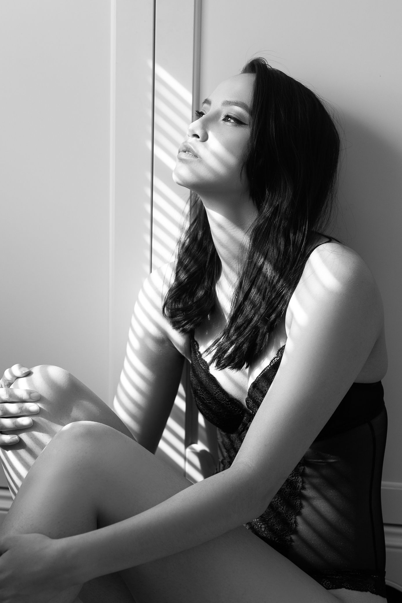 A dark-haired woman in a lace slip sits by a window with dramatic shadow stripes in black and white.