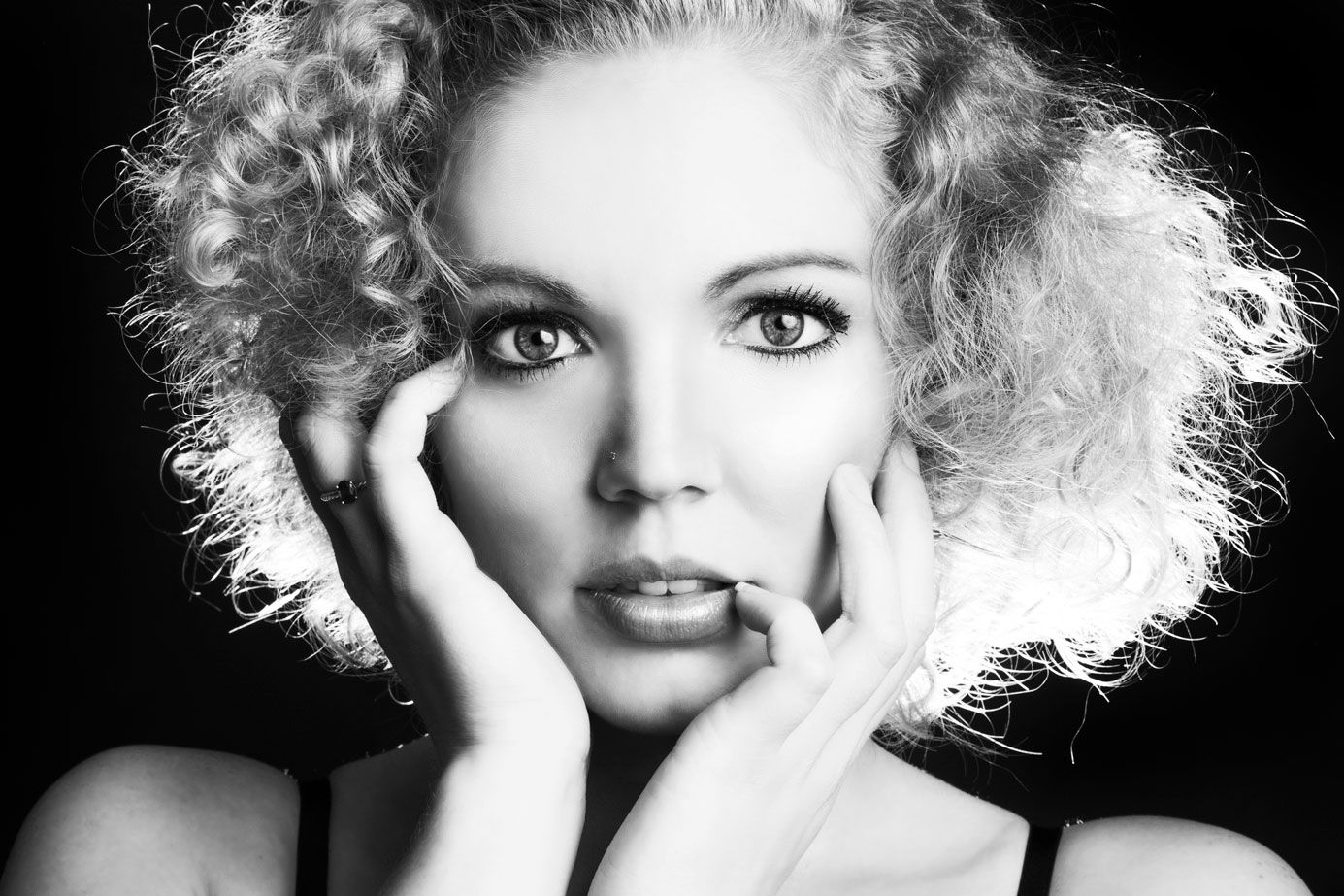 Black and white close-up portrait of a woman with curly blonde hair holding her face in her hands.
