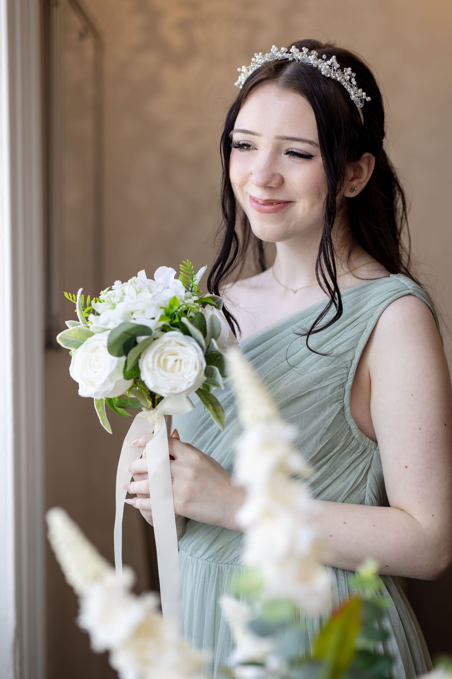 Smiling bridesmaid in sage green dress holding white rose bouquet with ribbon, wearing crystal tiara.
