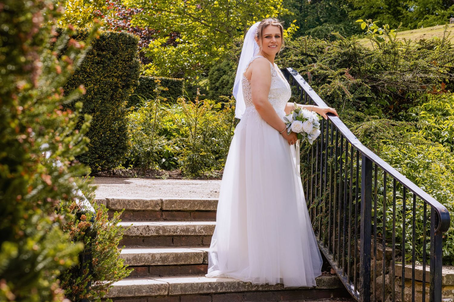 Bride in white wedding gown and veil holding bouquet on stone steps surrounded by lush green gardens.