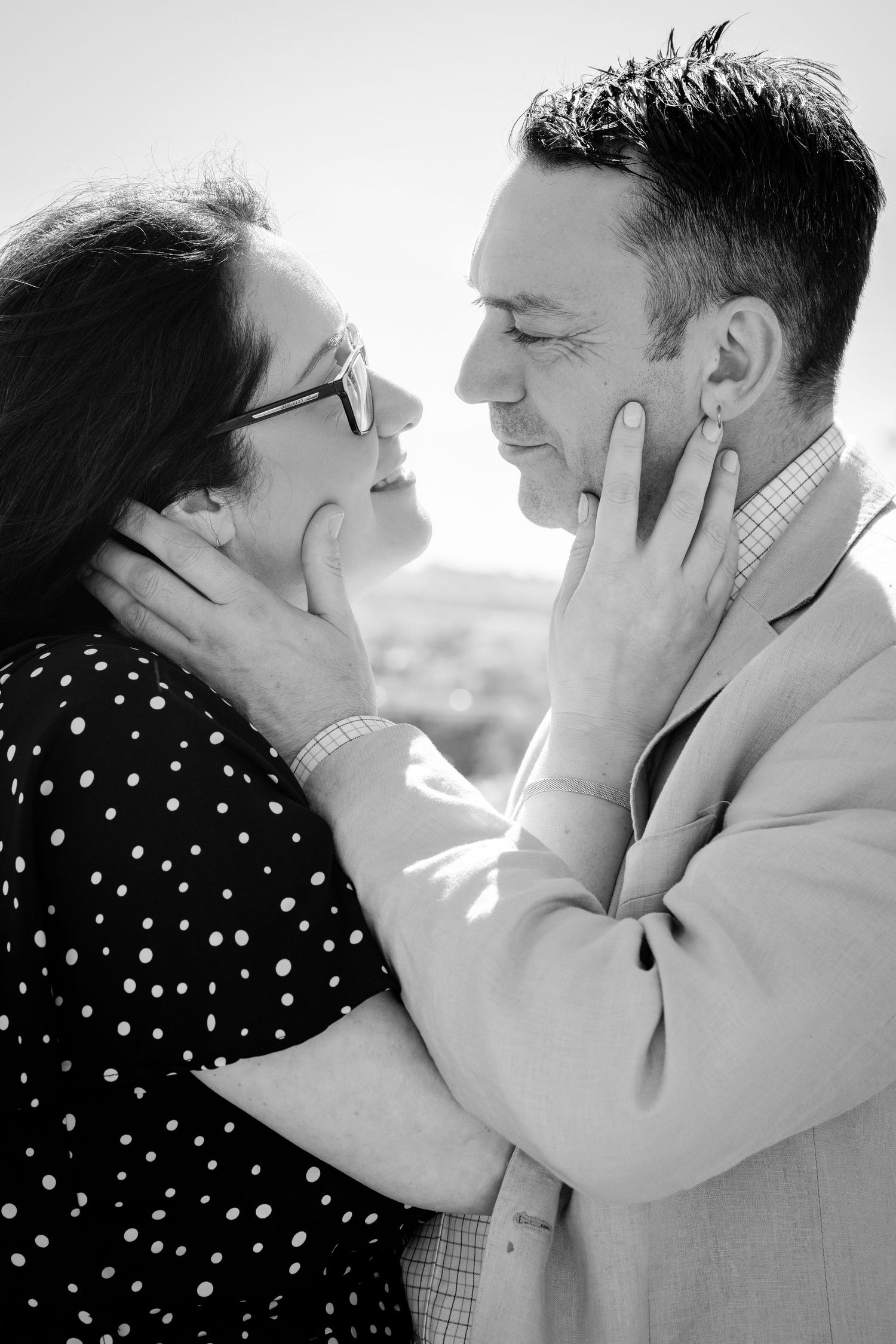Black and white photo of a loving couple holding each other's faces and smiling, dressed in polka dot and blazer.