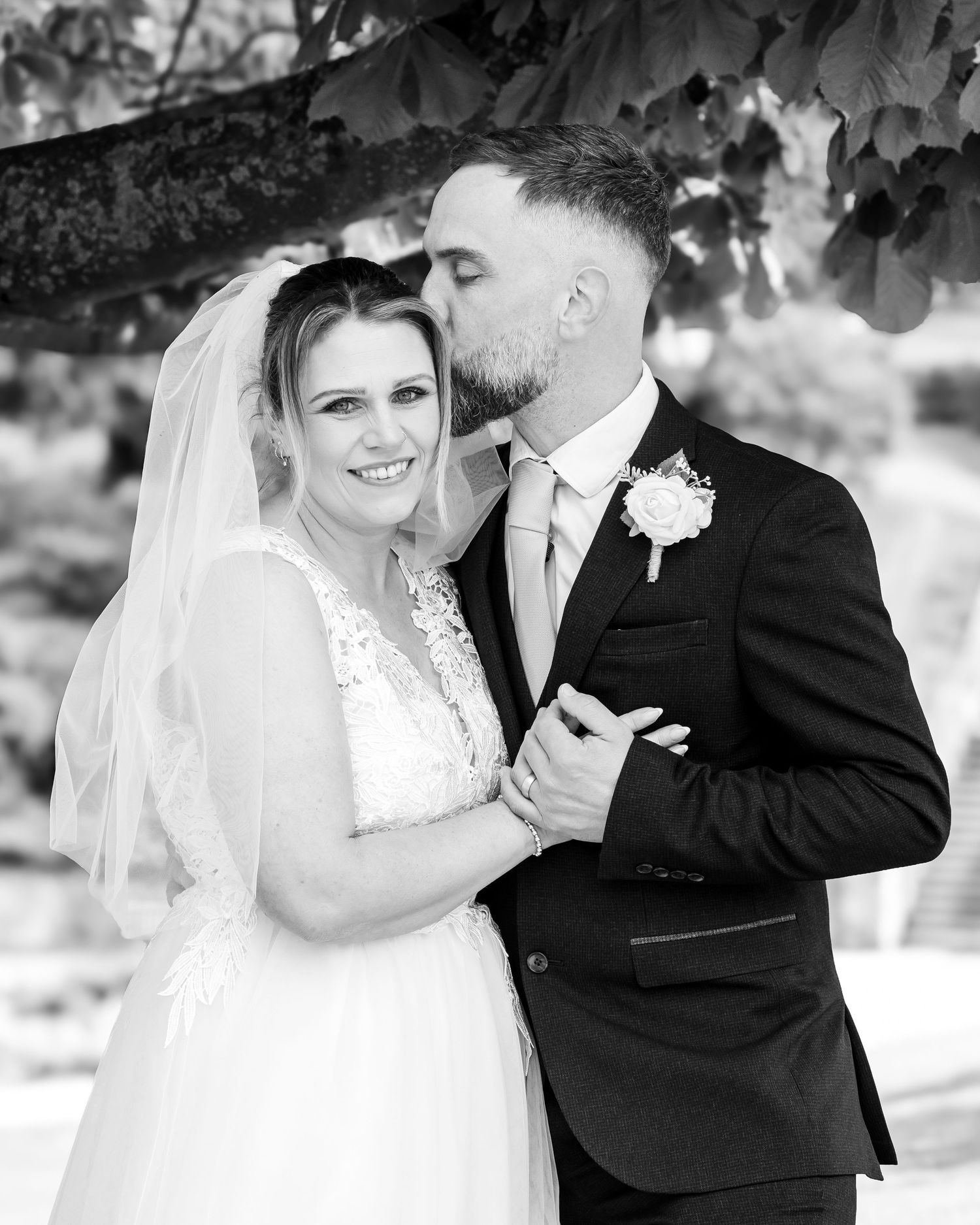 Black and white wedding photo of bride in lace gown and veil smiling as groom in suit kisses her cheek.