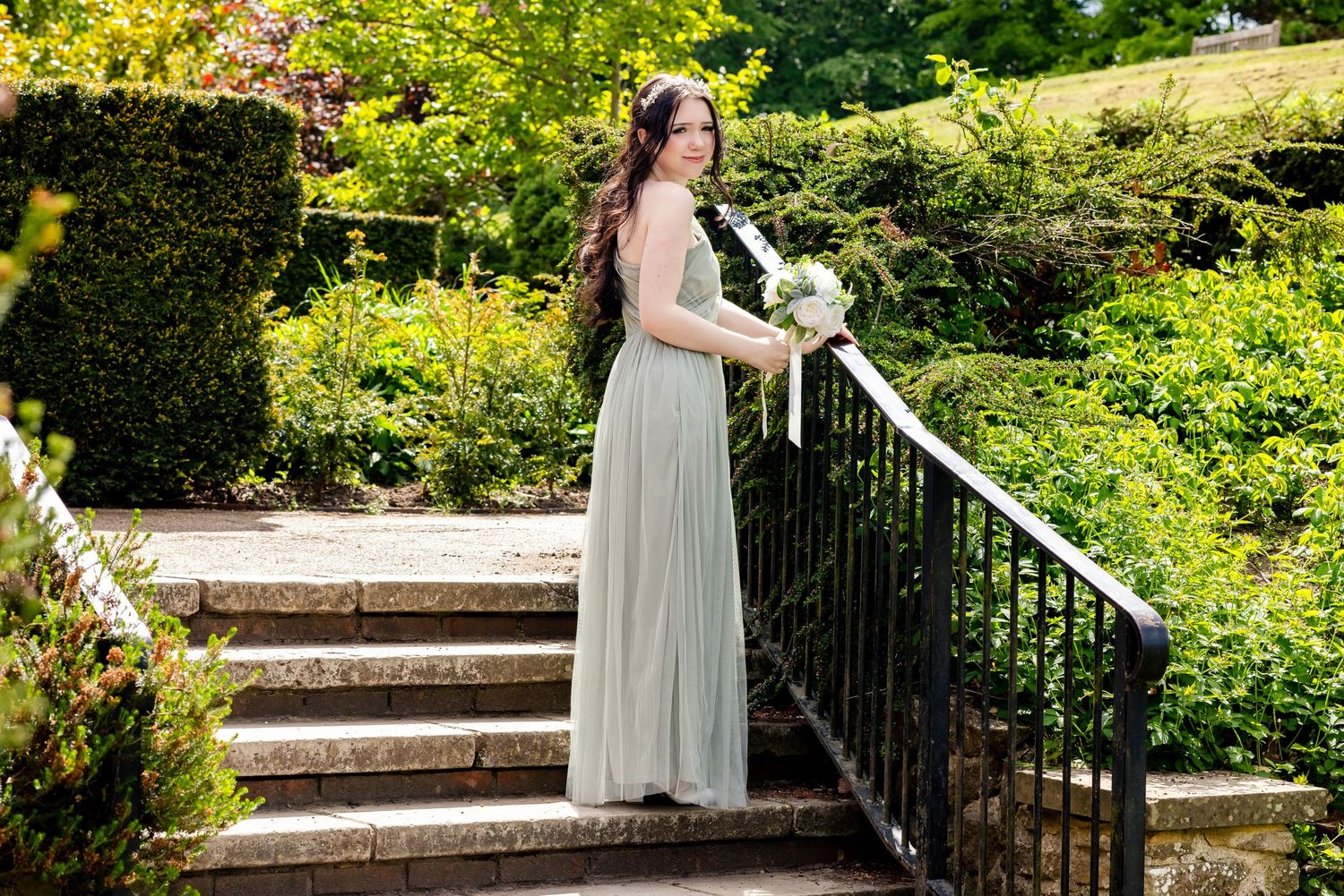 Elegant bridesmaid in sage green gown holding bouquet on stone steps surrounded by lush garden greenery.