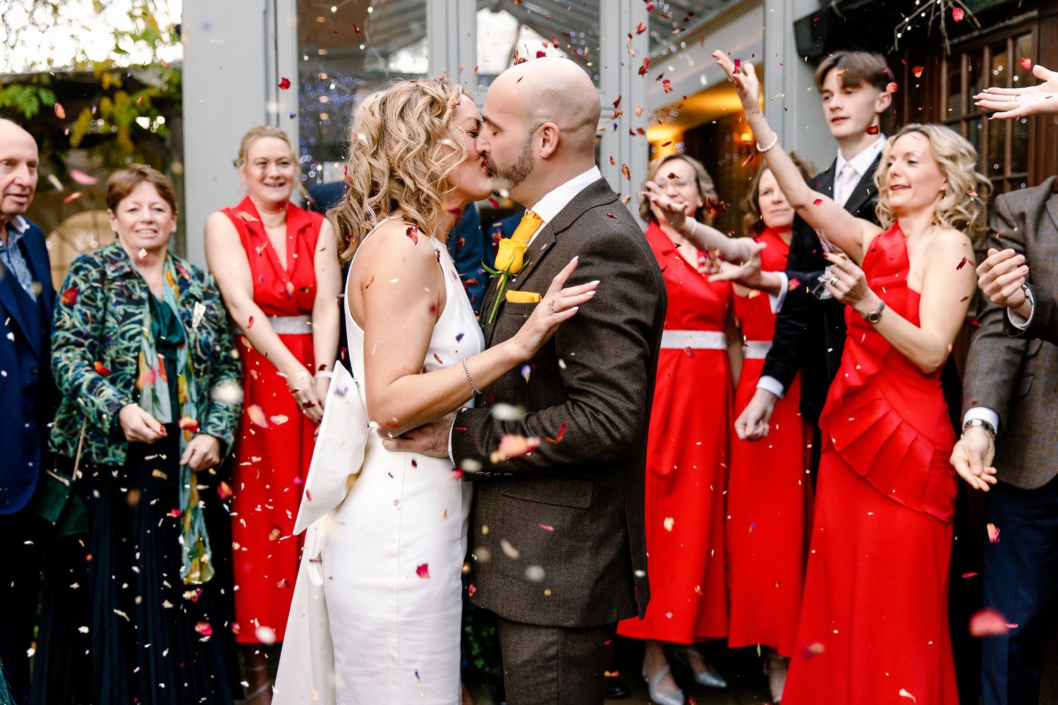 Bride and groom share first kiss surrounded by colorful confetti and cheering wedding guests.