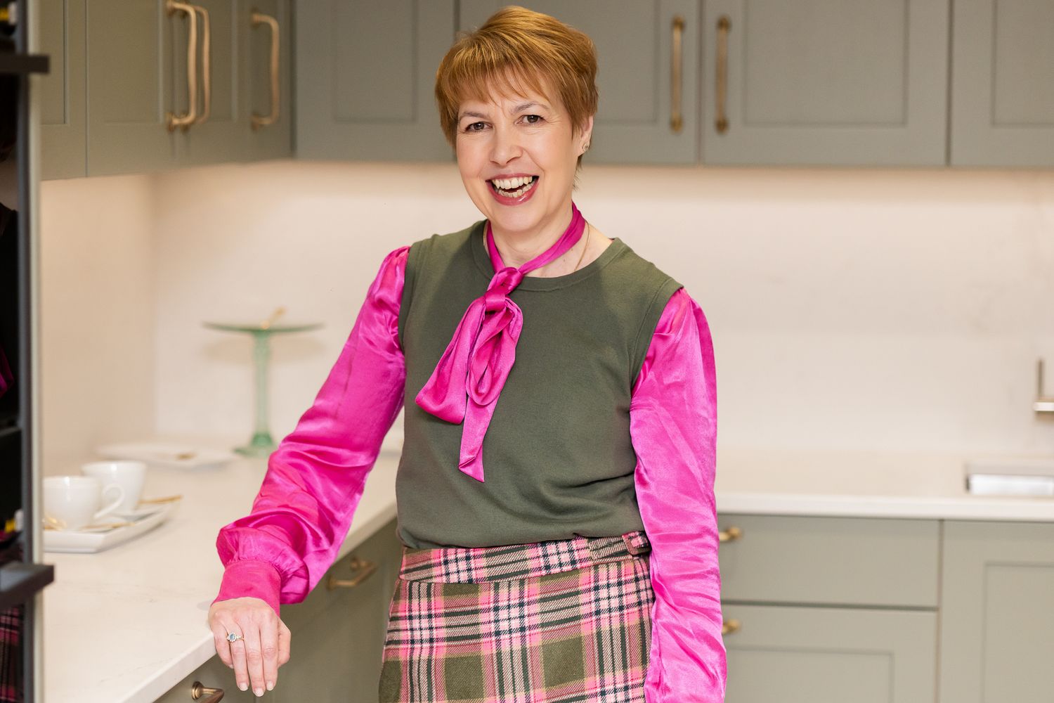 Woman in pink and green outfit smiling in a sage green kitchen.