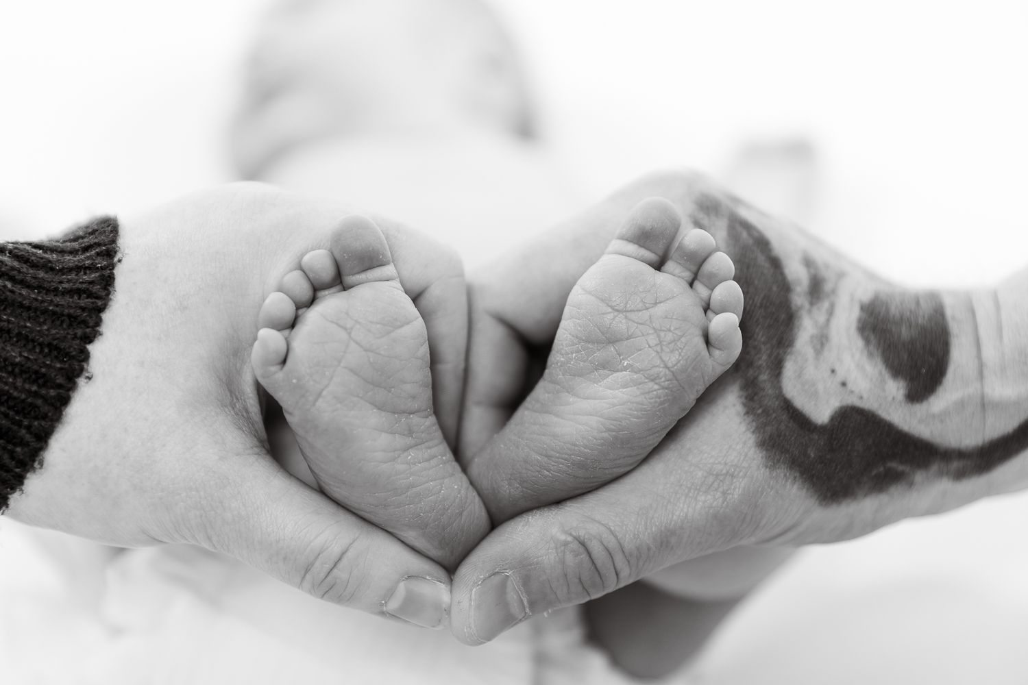 Parents forming a heart shape with their hands around their newborn babys tiny feet in black and white.