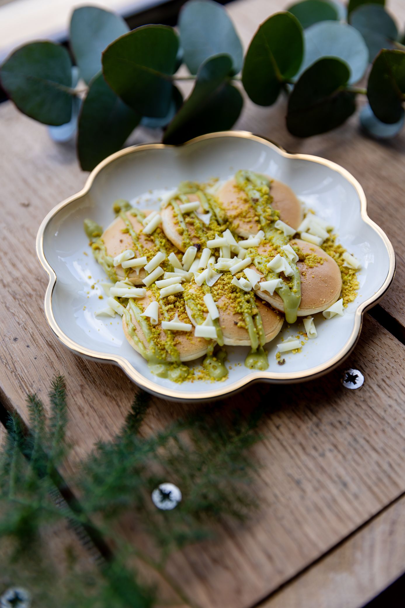 Overhead view of mini pancakes topped with pistachio cream and white chocolate on a decorative plate.