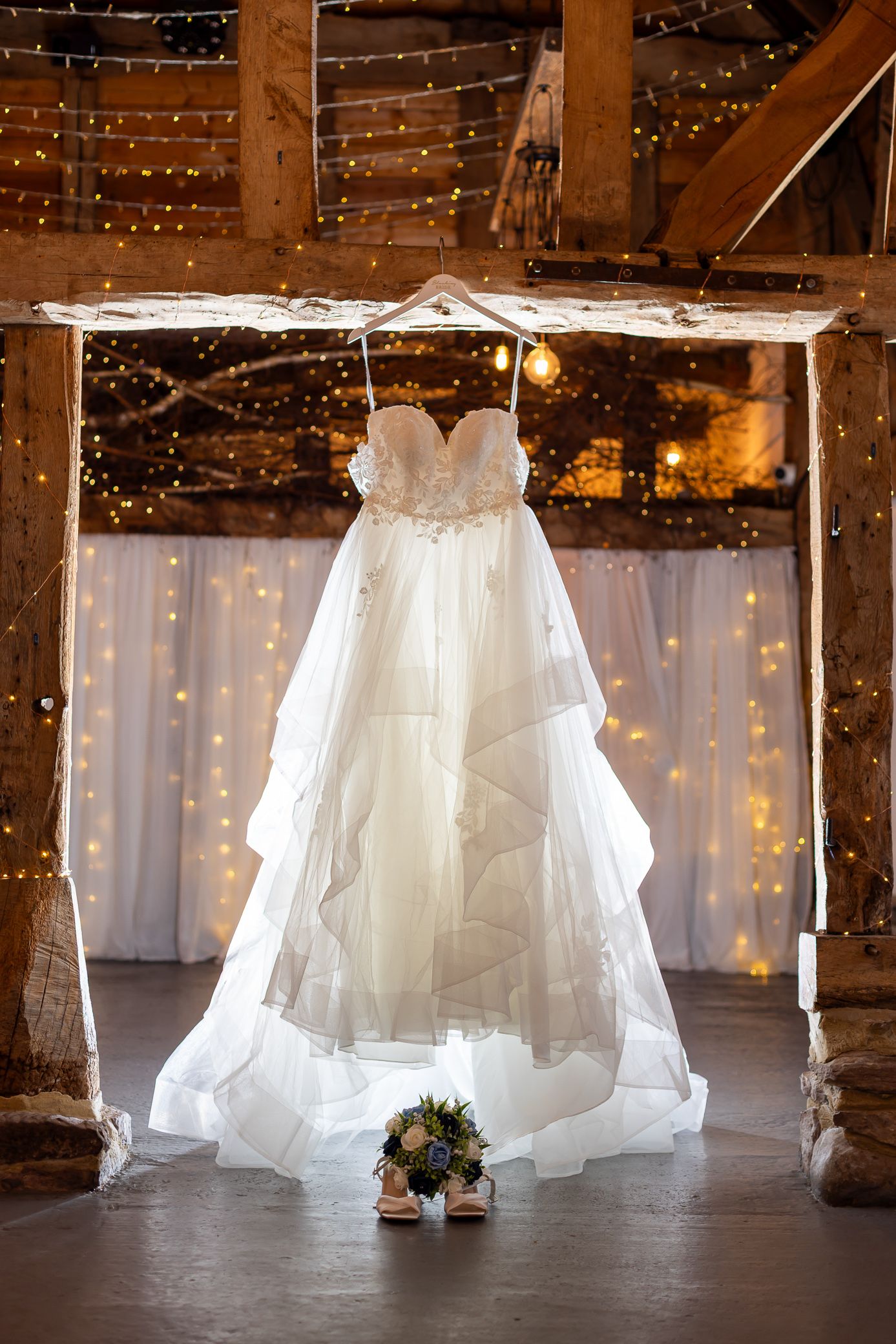 Strapless layered wedding gown hangs in a rustic barn adorned with fairy lights and white draping.