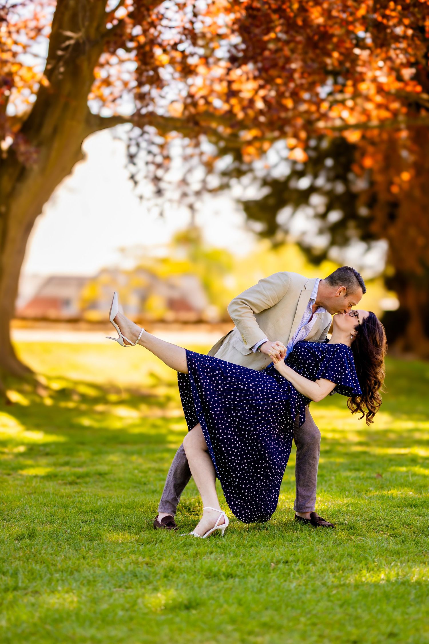 Couple dancing and kissing in a sunny park, woman in blue polka dot dress dipped by man in tan suit, autumn trees behind.