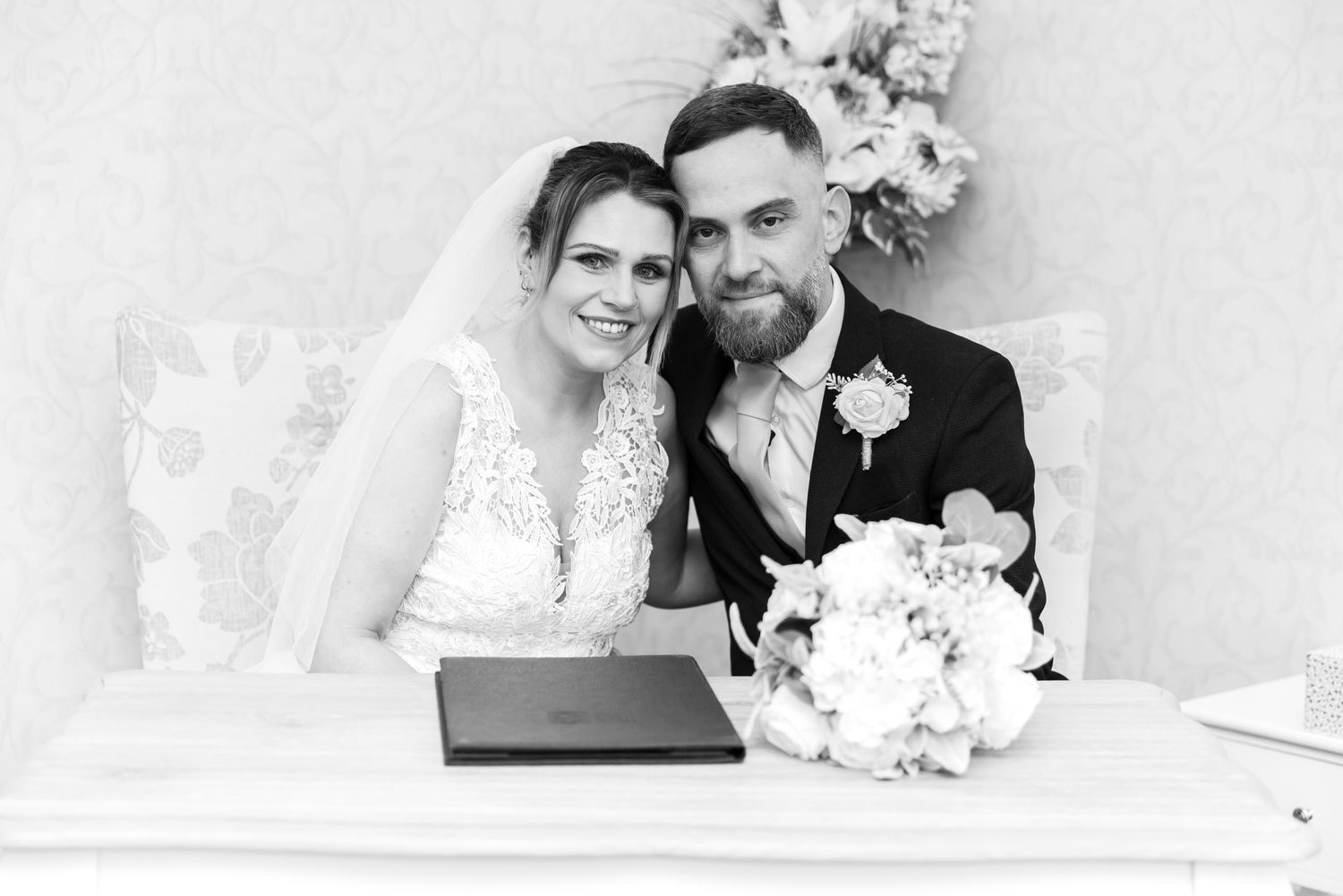 Black and white wedding photo of smiling bride and groom seated at signing table with bouquet.