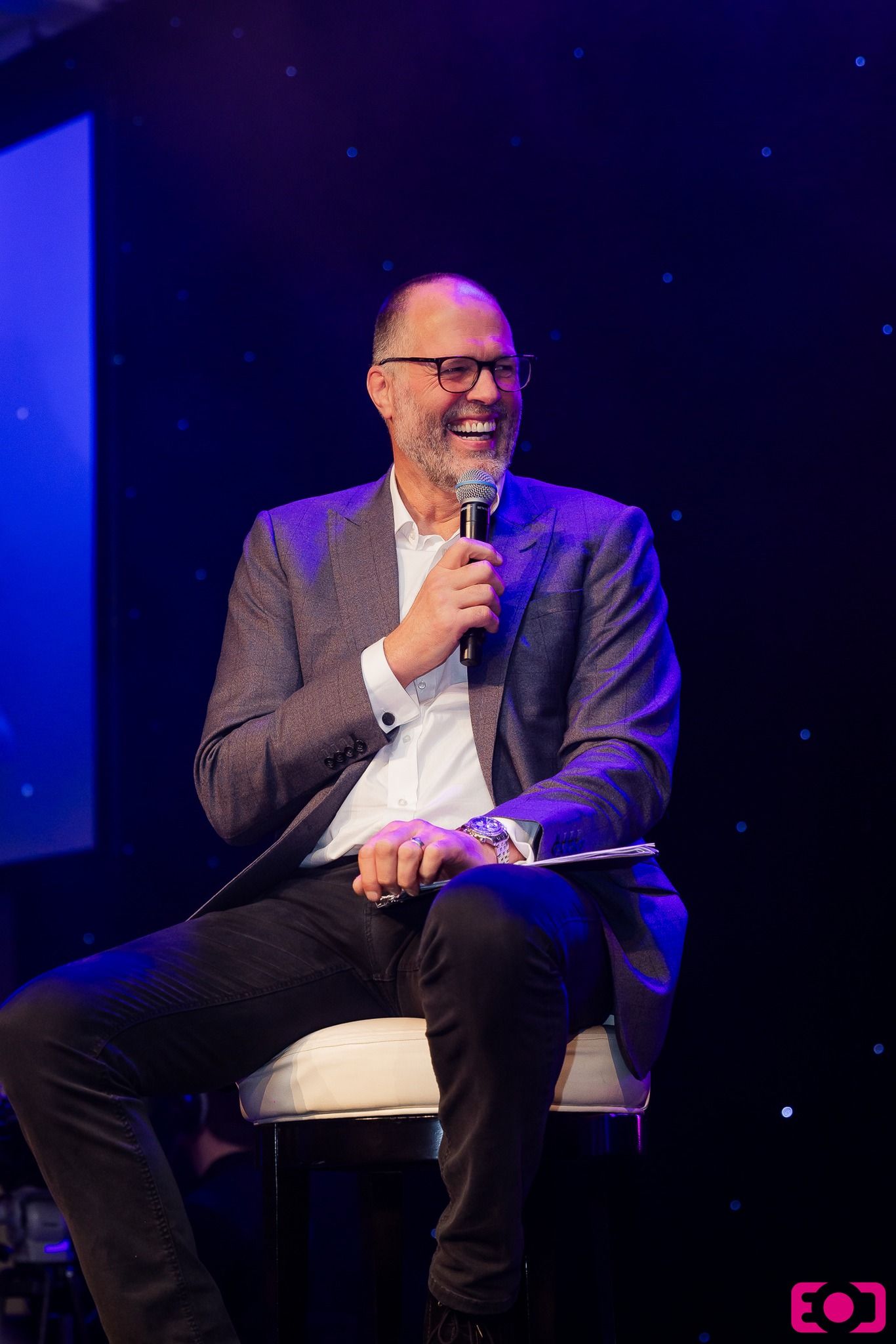 Smiling man in gray blazer holds microphone while seated on stage at an event.