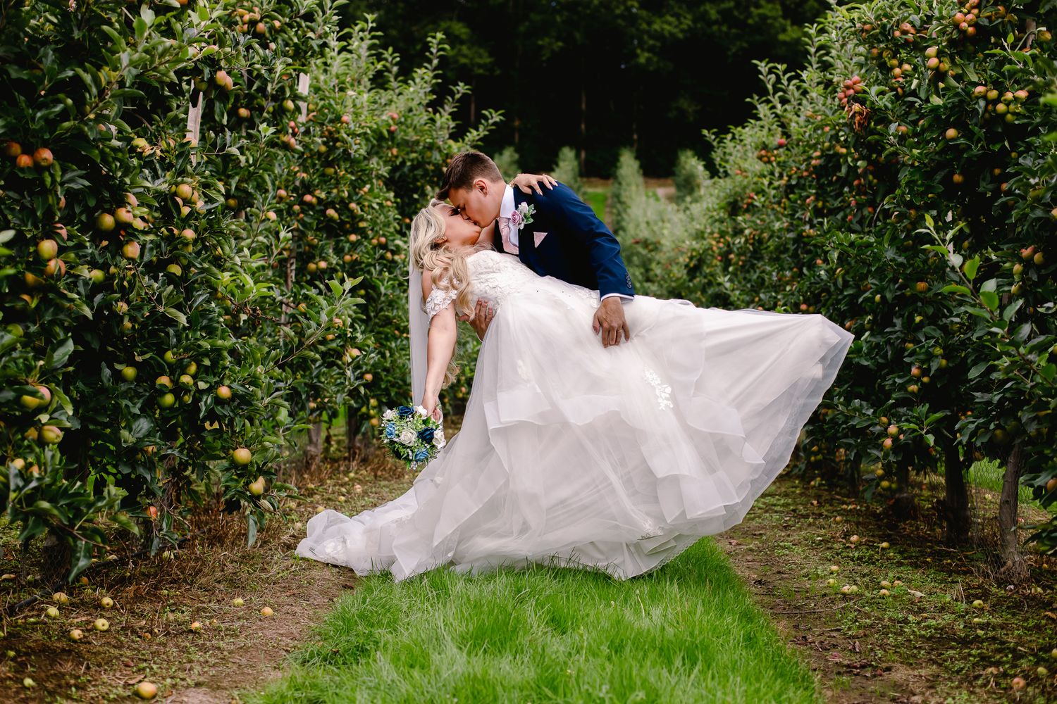 Groom lifts bride in a flowing white gown for a romantic kiss in a lush apple orchard.