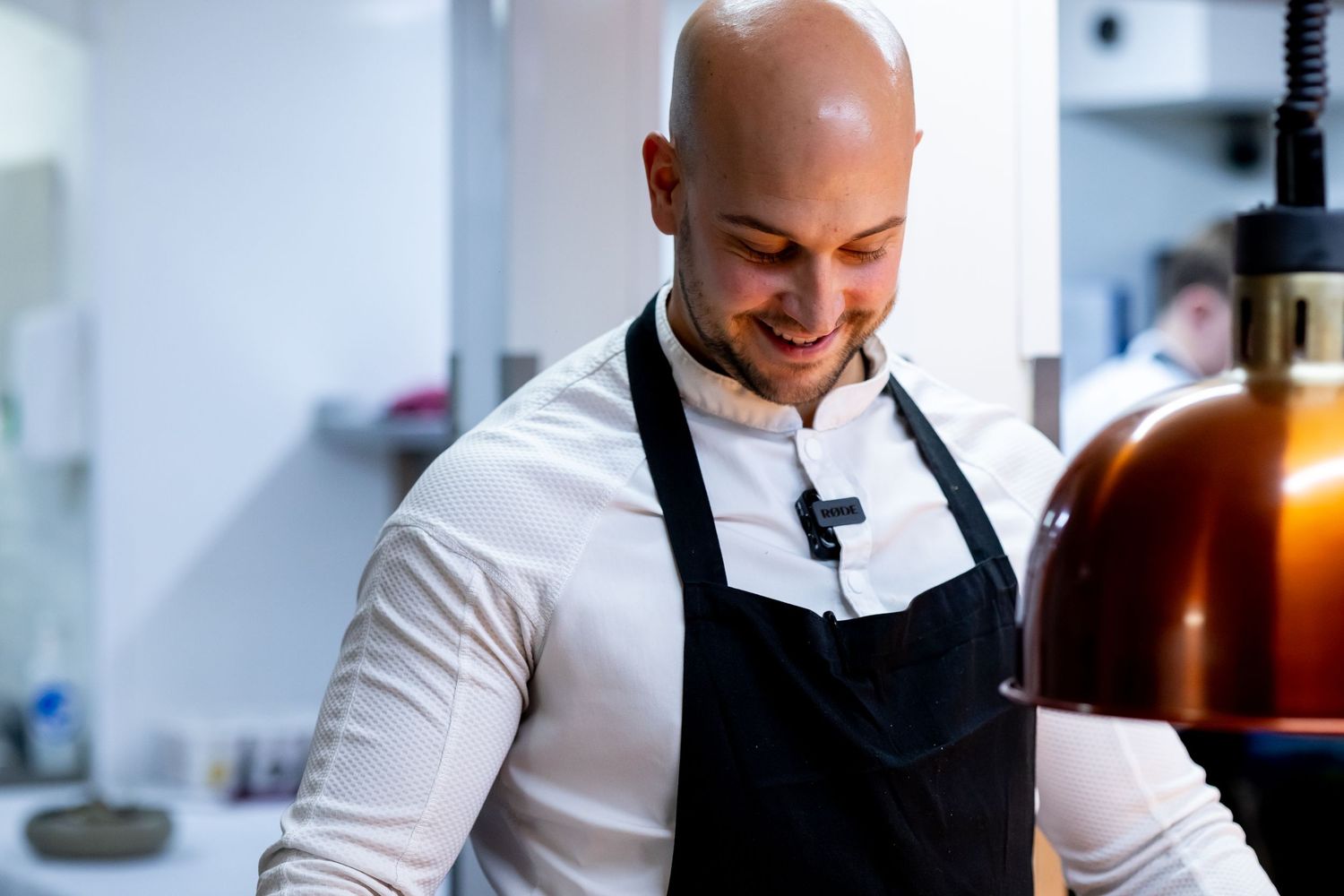 Smiling bald male chef in black apron and white shirt working in a professional kitchen.