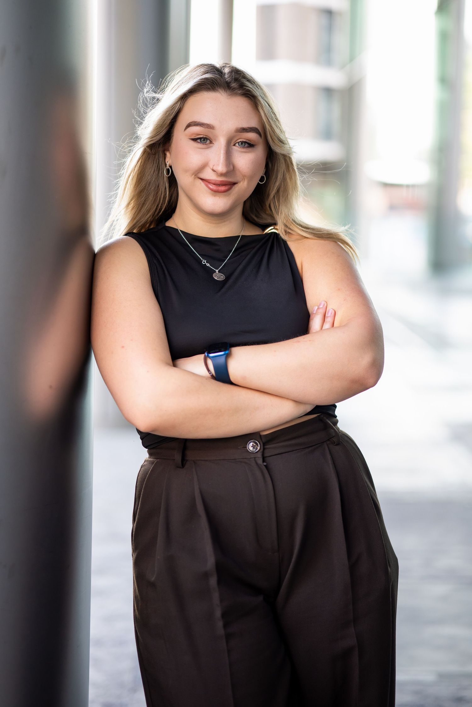 Young blonde woman in black top and brown trousers stands confidently with arms crossed, leaning against a pillar outdoors.