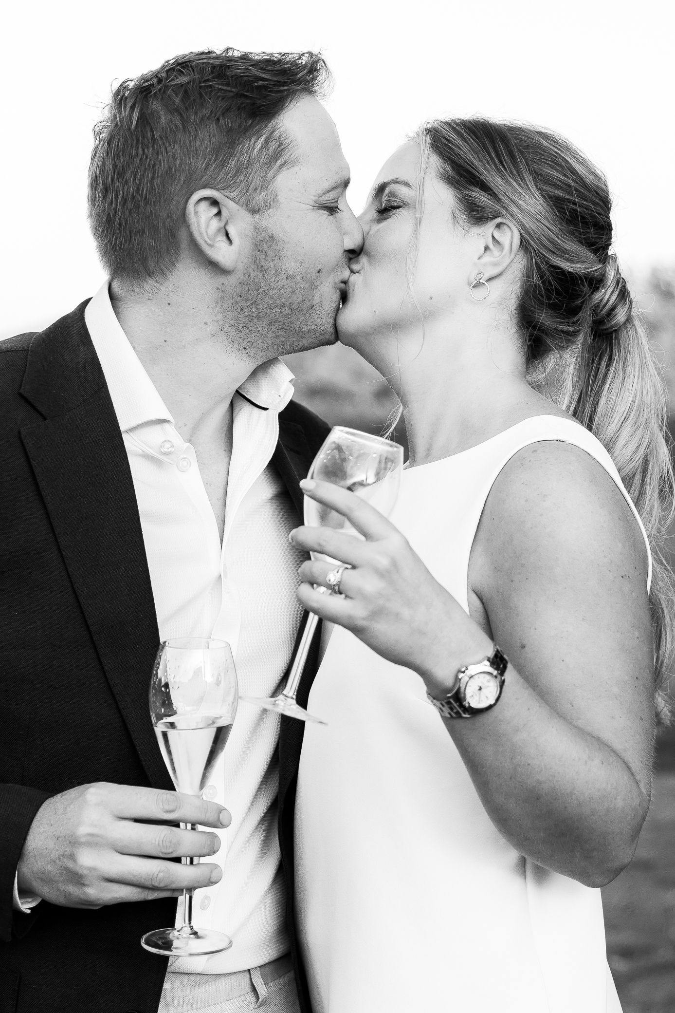 Black and white photo of couple kissing while holding champagne glasses at their wedding celebration.
