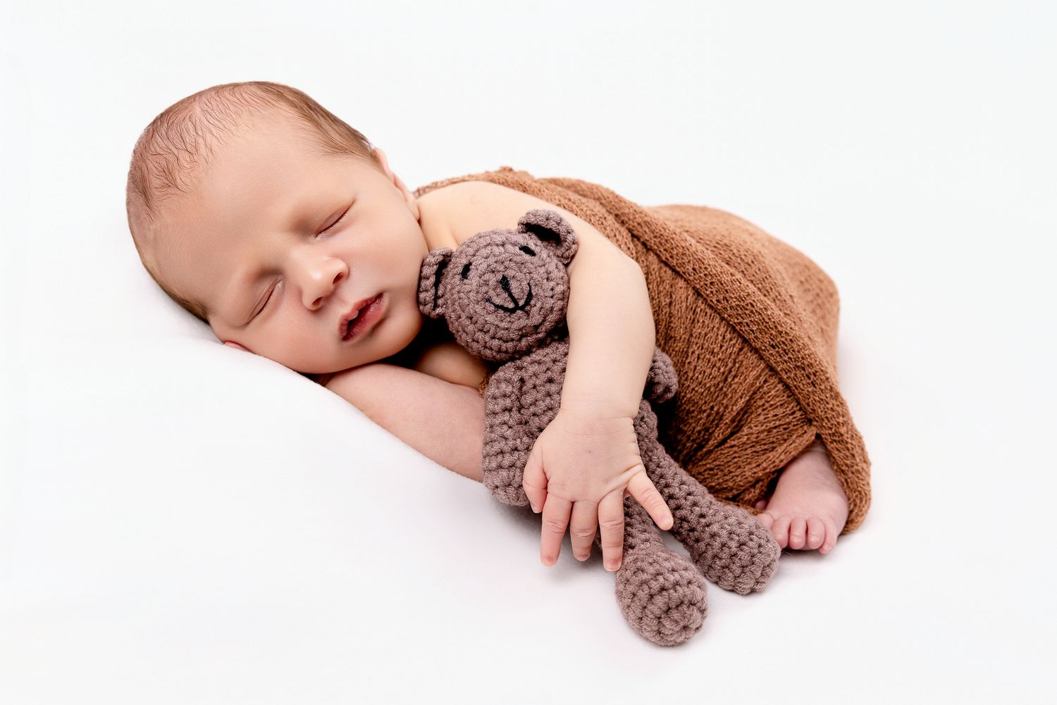 Sleeping newborn baby in brown knit outfit holding a small crocheted teddy bear.
