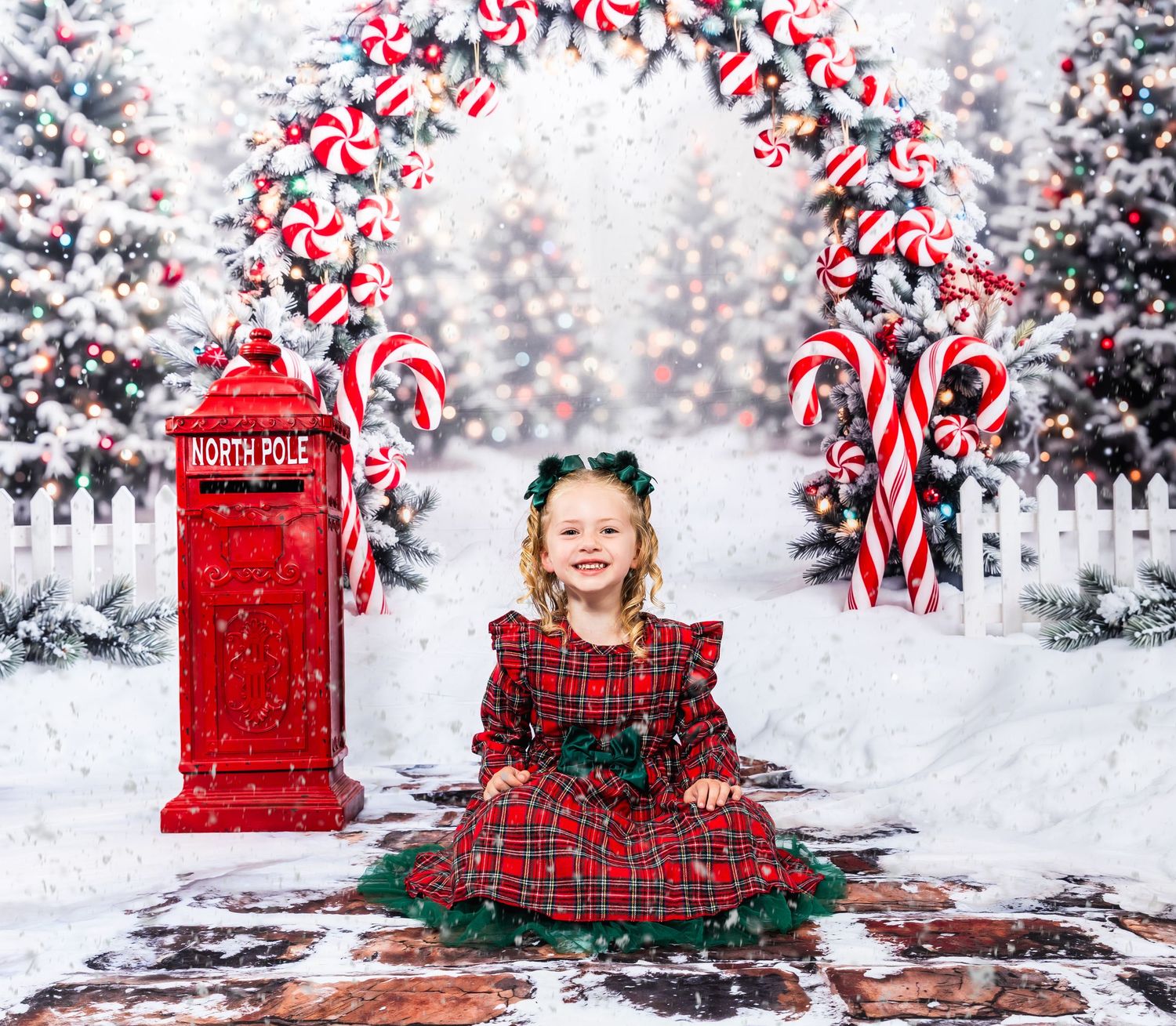 A young girl in a red plaid dress sits before a festive North Pole backdrop with candy canes and snowy Christmas trees.