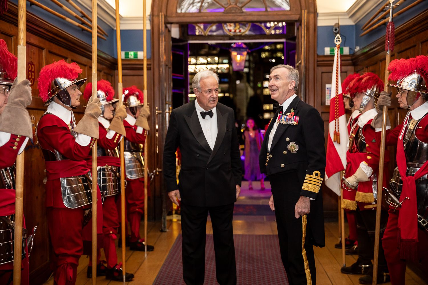 Two formally dressed men walking between ceremonial guards in red uniforms at an elegant evening event.