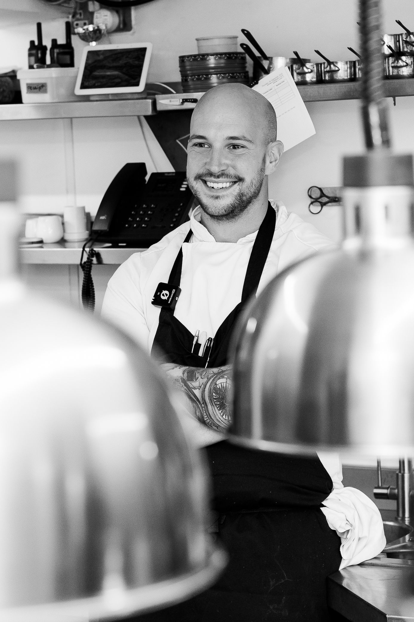 Smiling bald chef in black apron posing in a professional kitchen, black and white photo.
