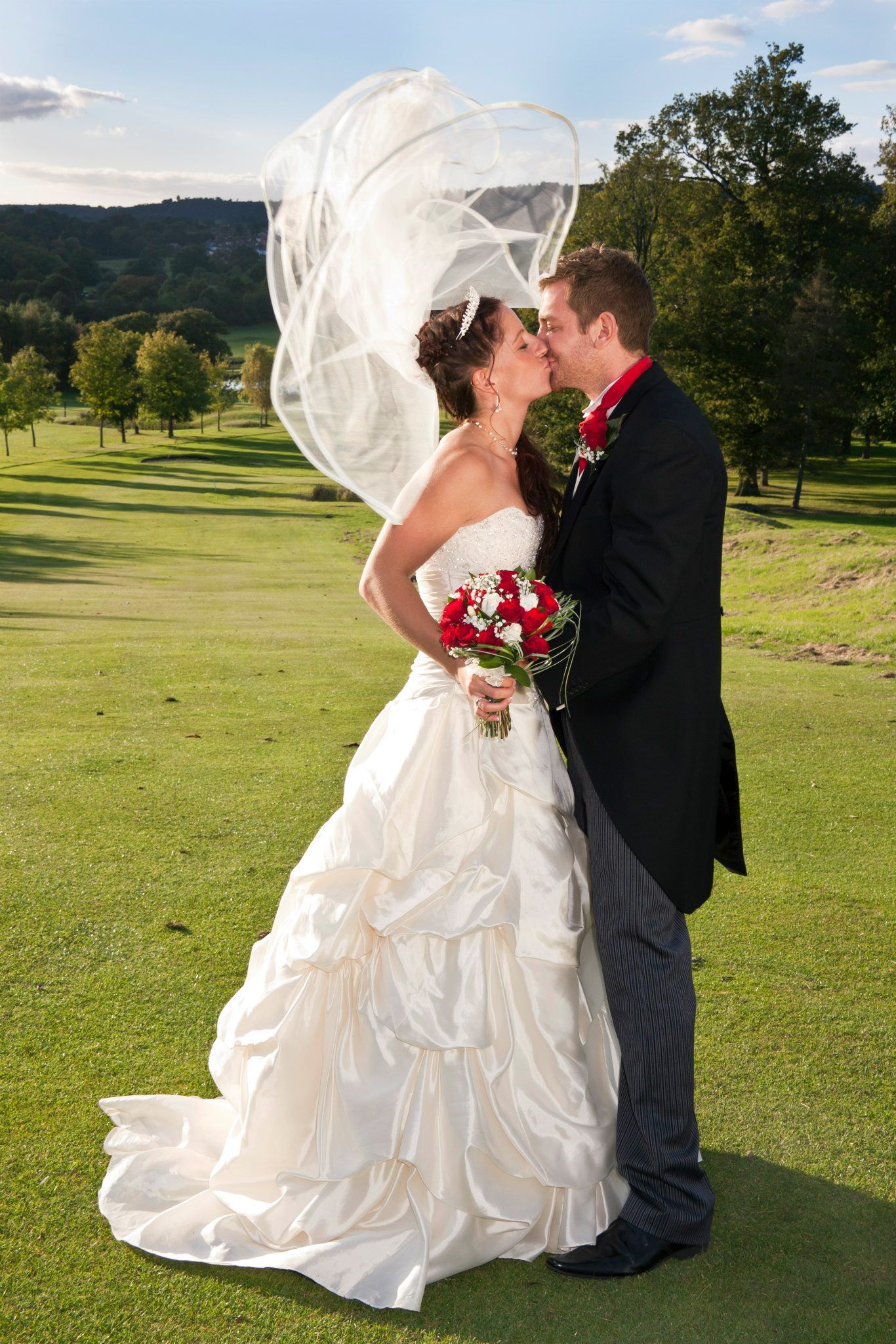 Bride and groom kissing on golf course with veil flowing in wind, romantic outdoor wedding photography.