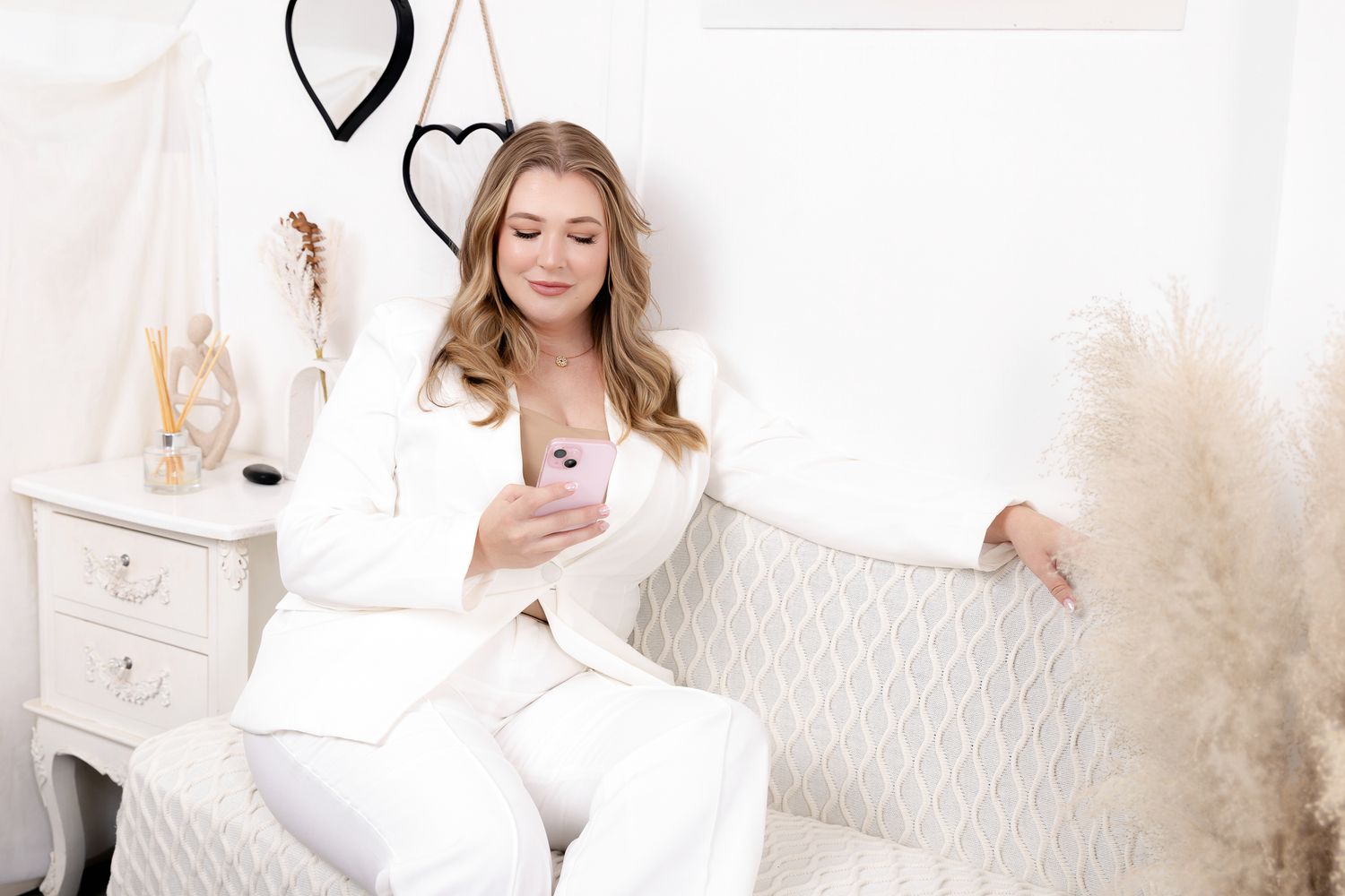 Blonde woman in white suit using a pink smartphone in a bright white room.