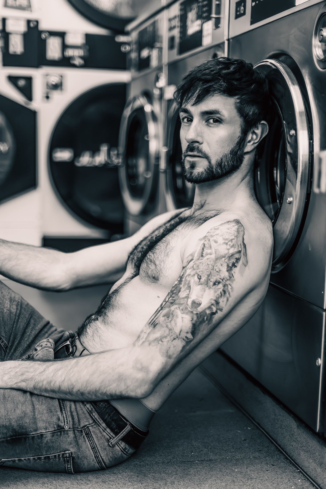 A tattooed shirtless man in jeans sits against laundry machines in a black and white photo.
