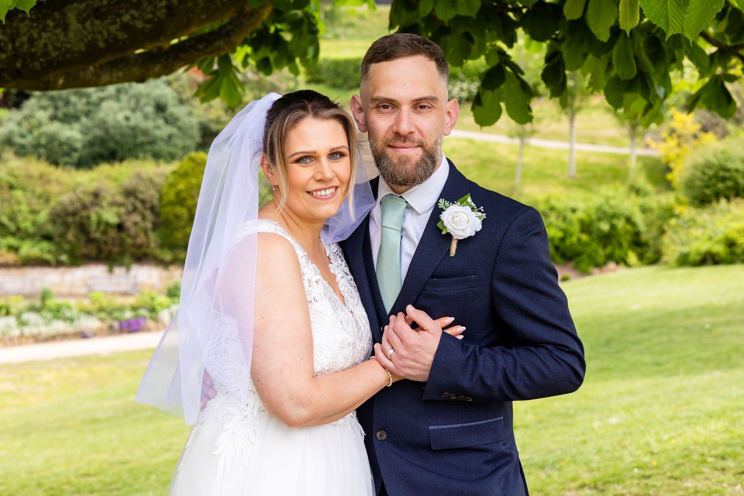 Happy bride and groom posing outdoors in a lush green garden on their wedding day.