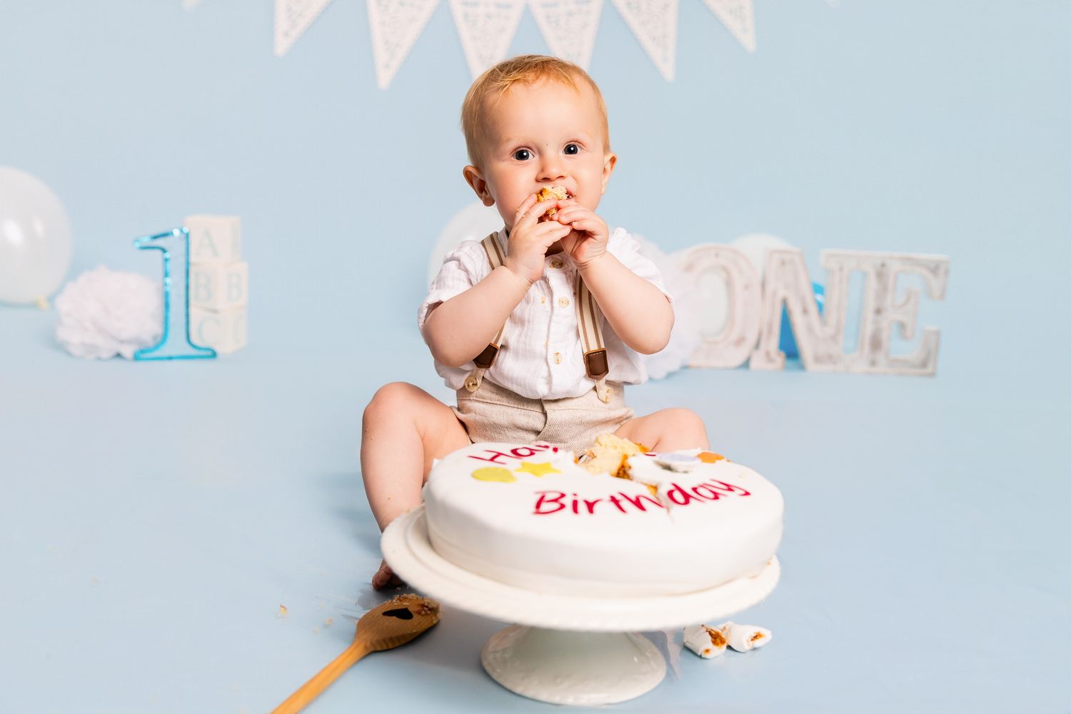 Baby boy celebrating first birthday smashing white cake, sitting against light blue background with ONE decorations.