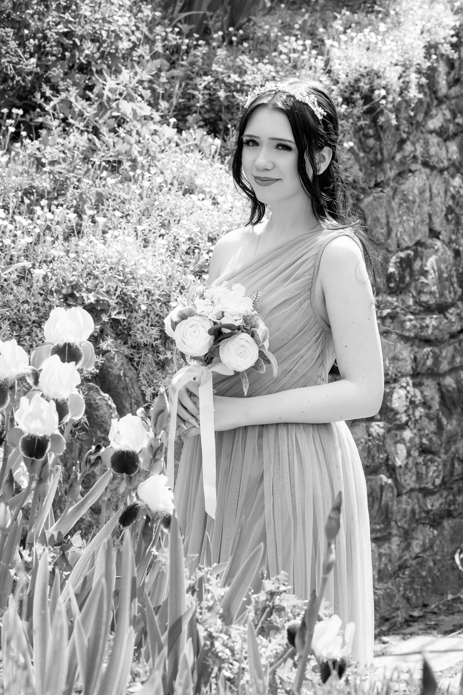Black and white photo of a bridesmaid in a flowing gown holding a bouquet of roses in a garden setting.