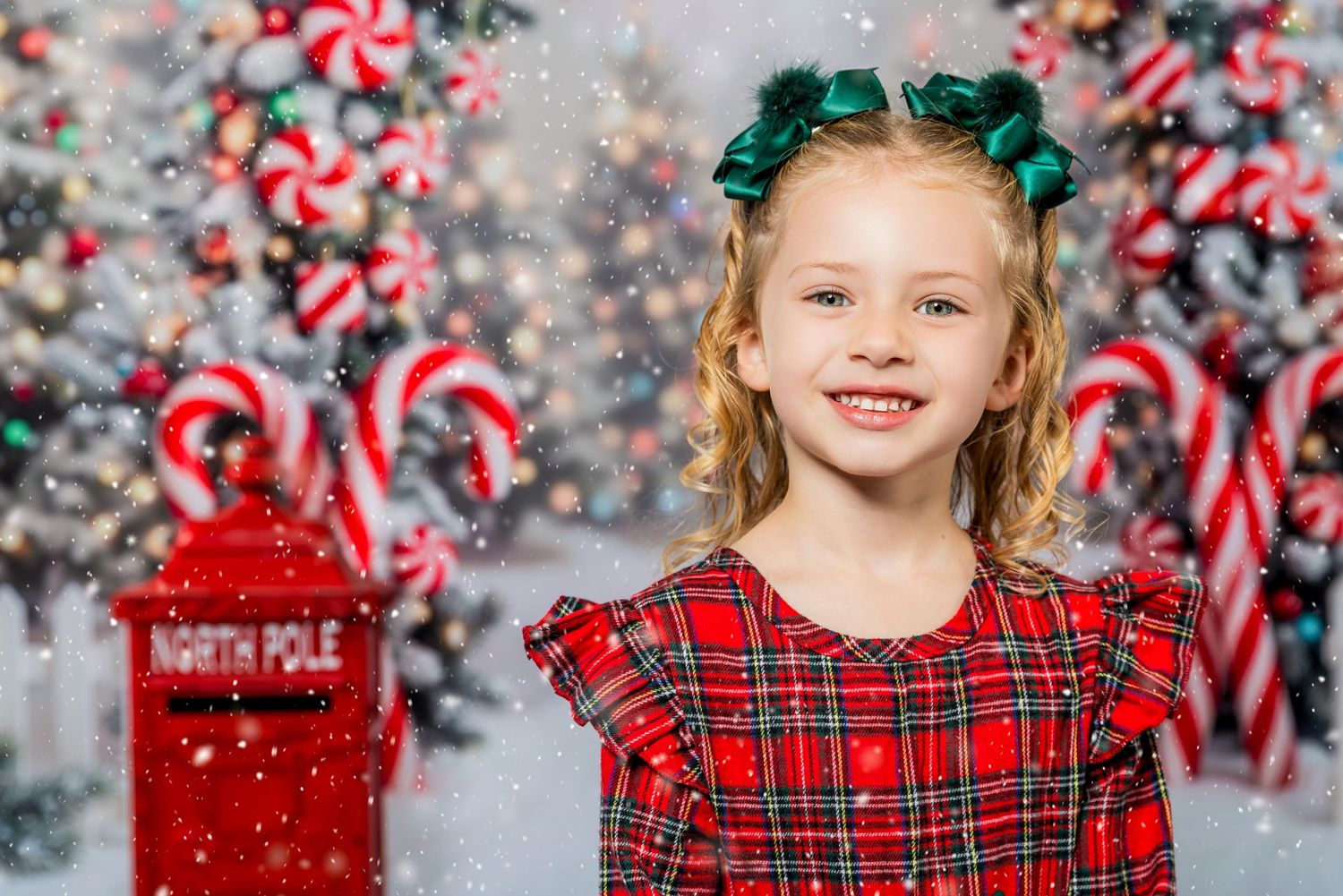 A young girl in a red plaid dress with green hair bow smiles in front of a festive North Pole Christmas backdrop.