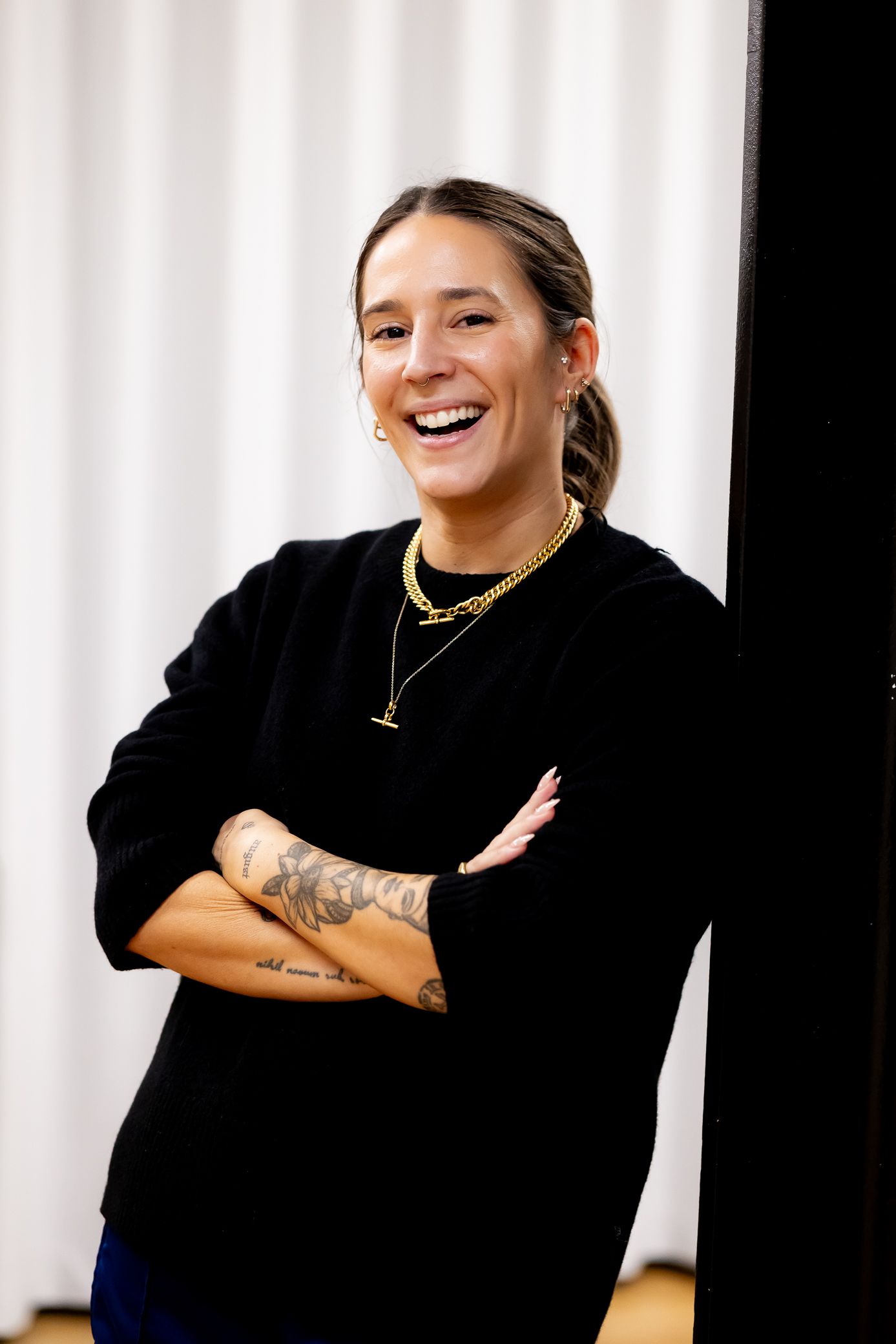 Laughing woman in black sweater with tattoos and gold necklaces standing with arms crossed against white curtain.