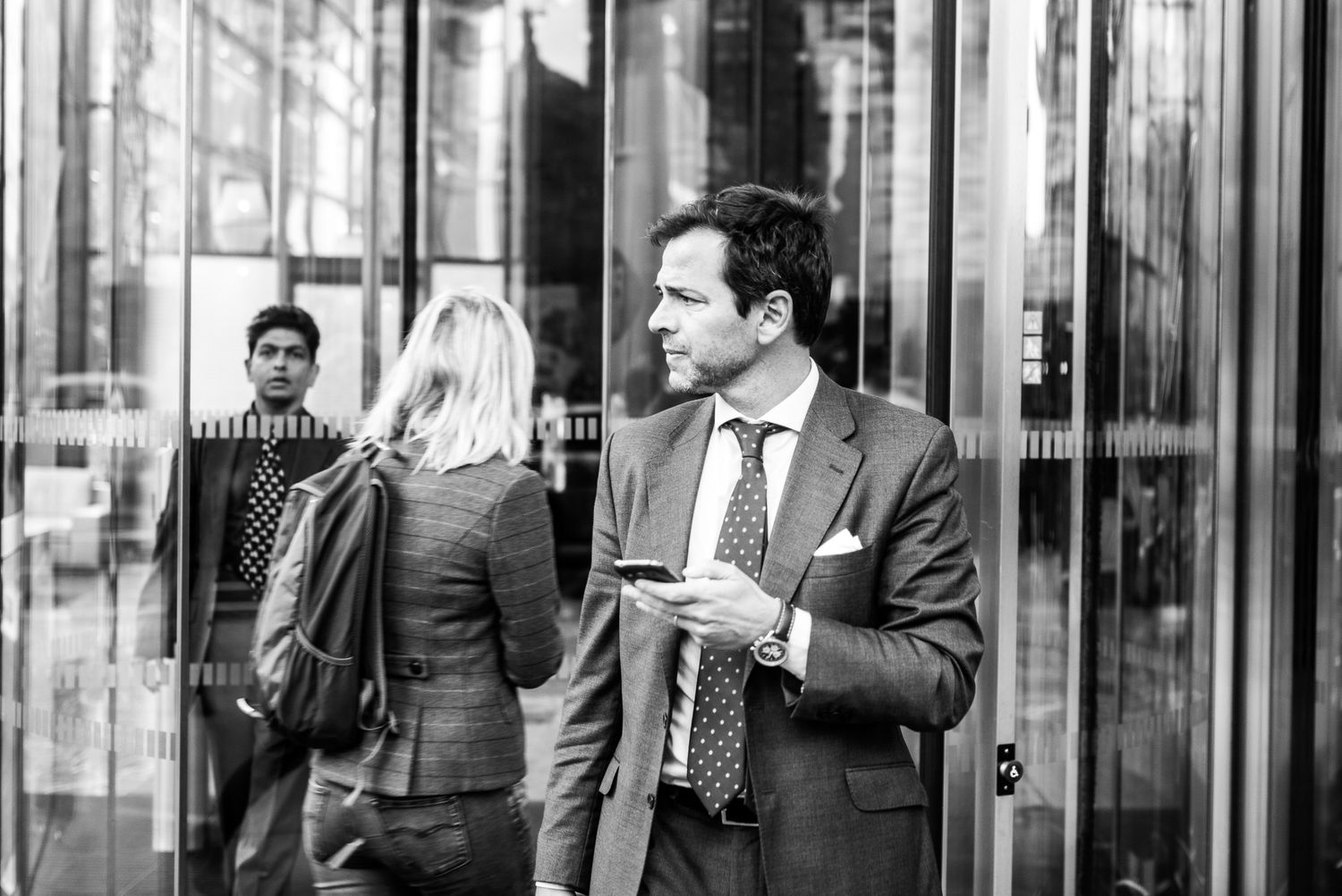 Businessman in suit checking phone outside revolving glass doors in black and white urban photo.