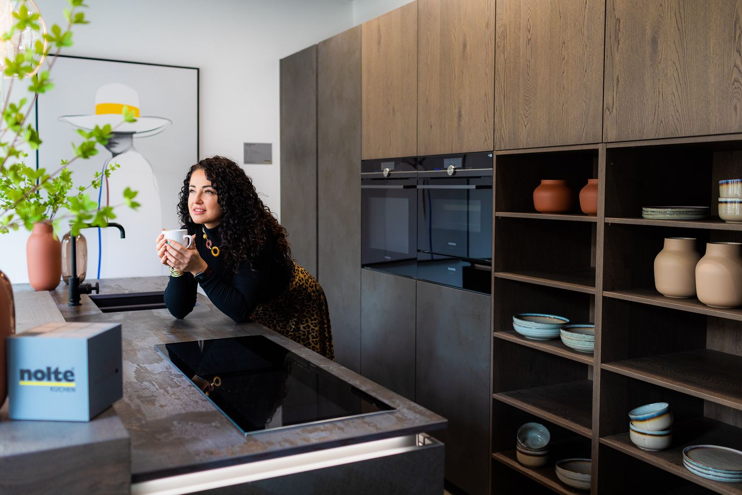 Woman relaxing at a Nolte kitchen island with open shelving displaying ceramics and a built-in oven.