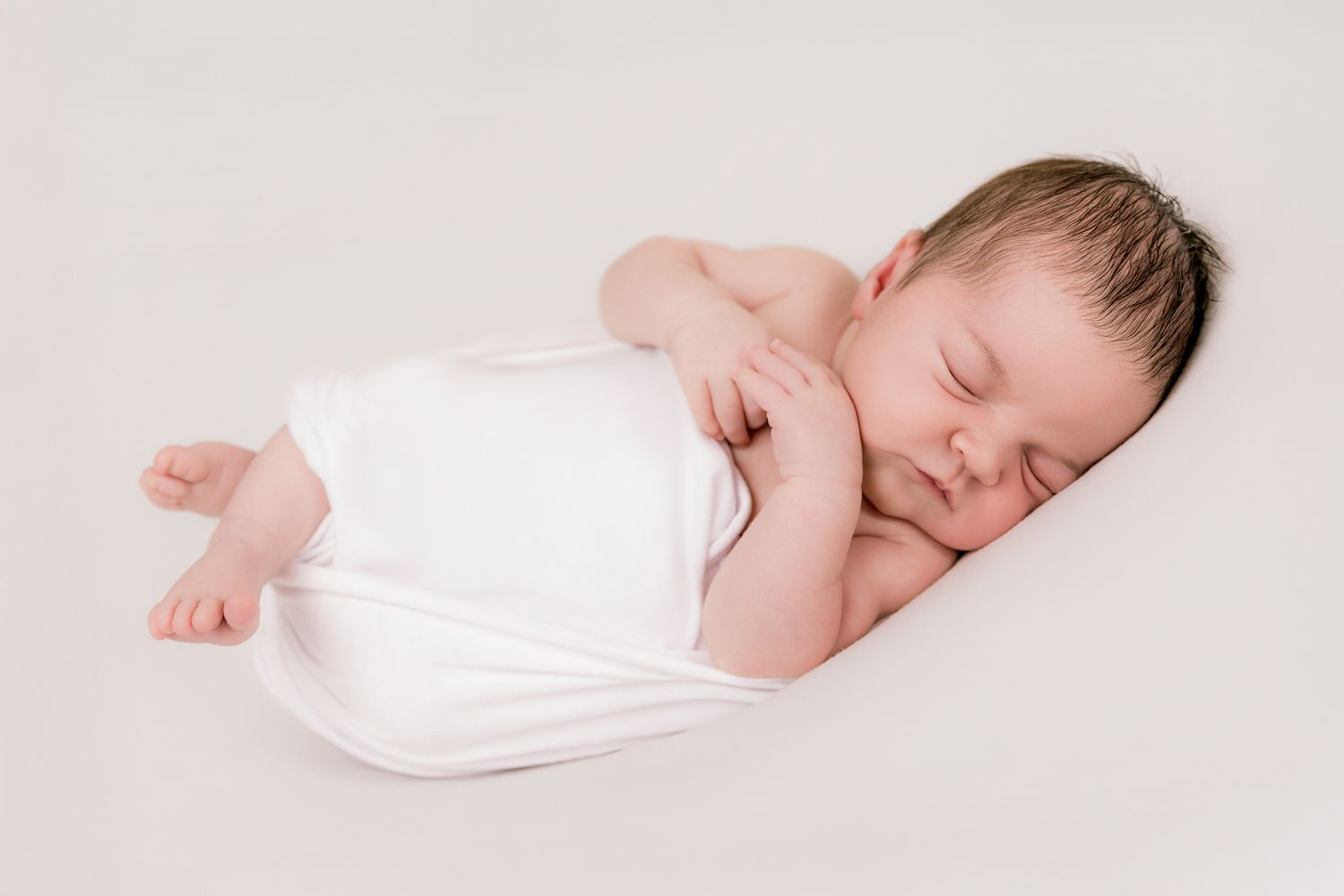 Peaceful sleeping newborn baby wrapped in white cloth, lying on soft white background.