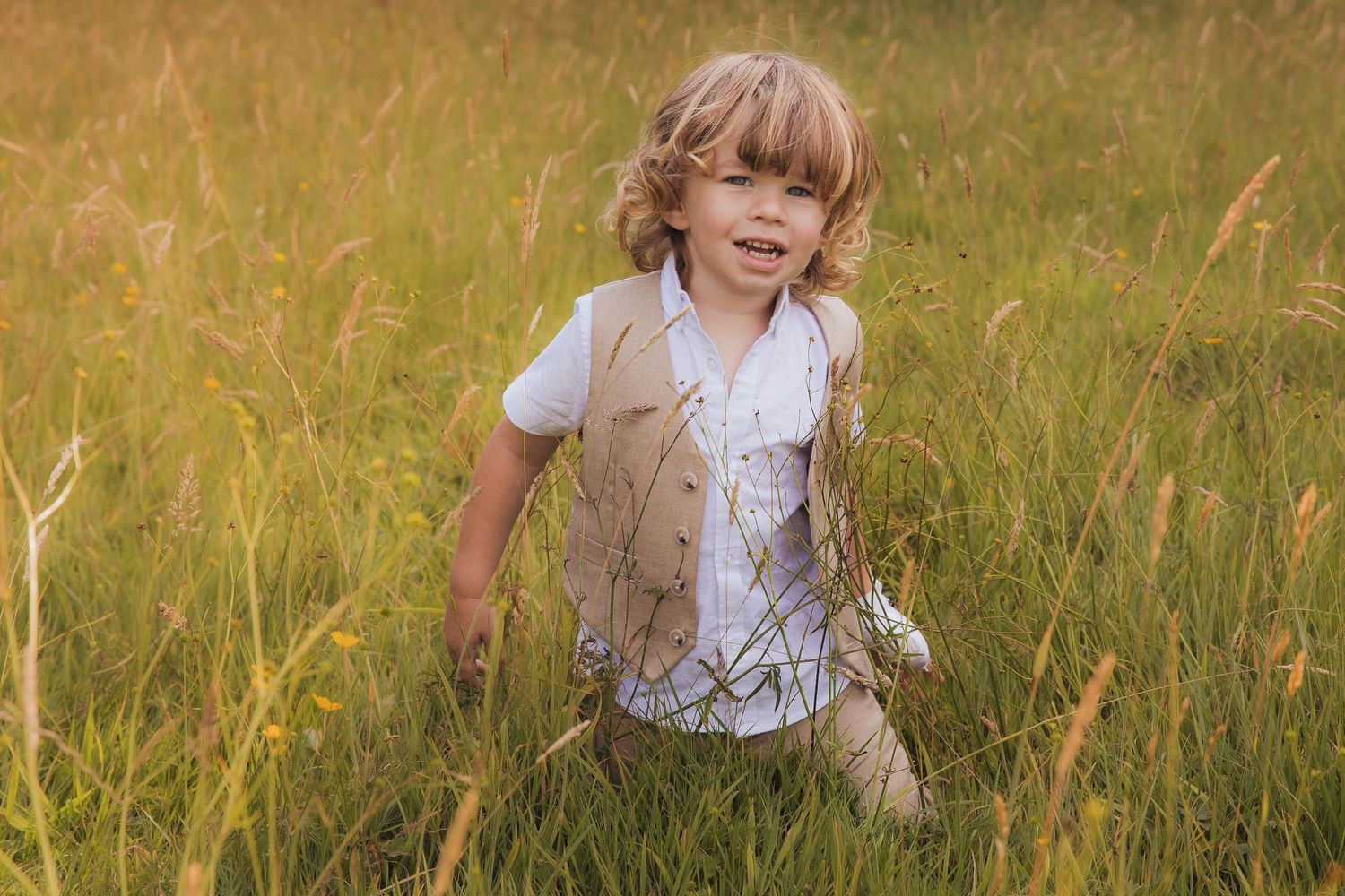 Young blond boy in a vest playing in tall golden grass at an outdoor wedding celebration.