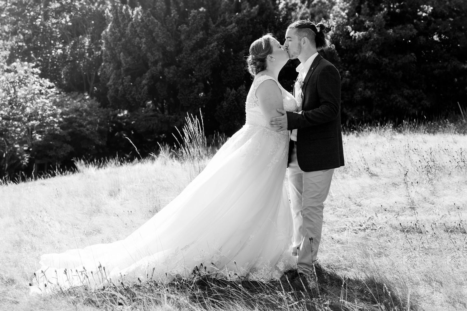 Black and white photo of bride and groom embracing in a grassy field with trees in the background.