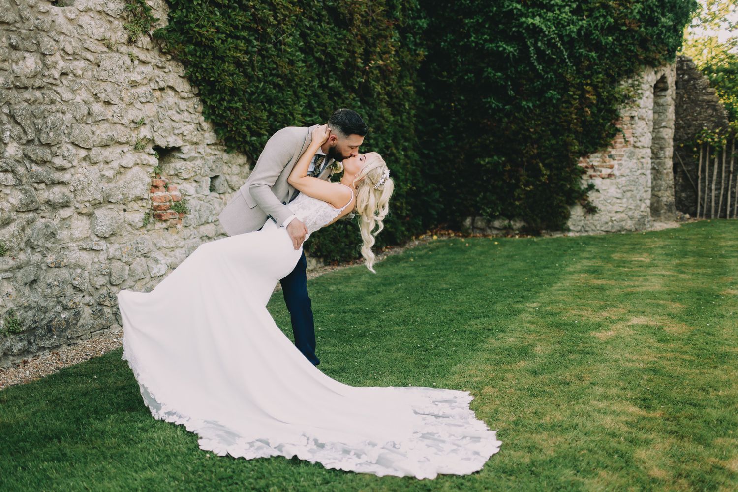 Groom dipping bride in stunning lace wedding dress against ivy-covered stone wall.