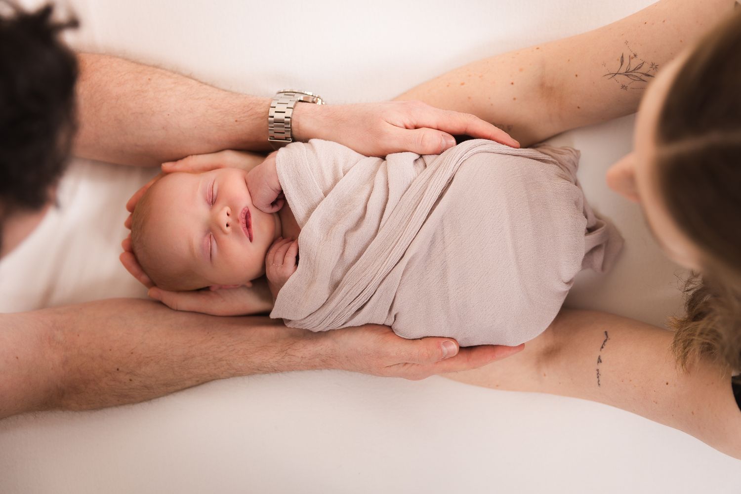 Parents gently cradling their sleeping swaddled newborn baby from above in a studio setting.